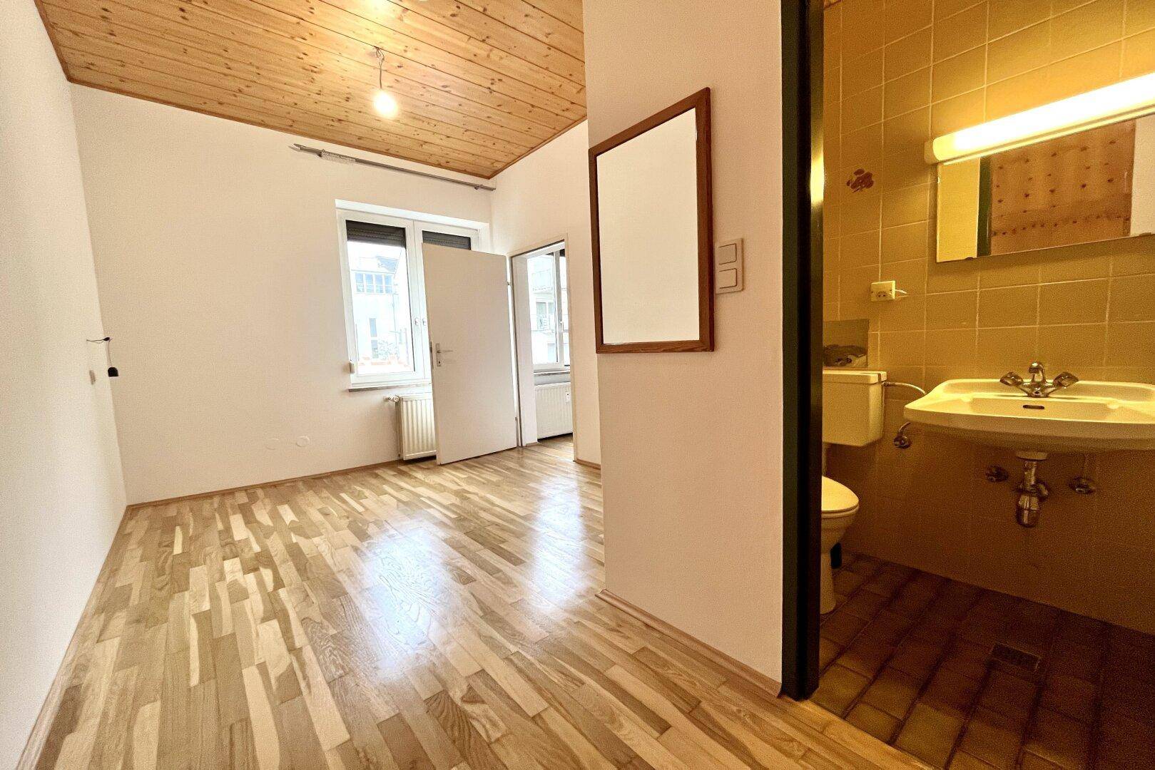 Heller Raum mit Holzbalkendecke und Parkettboden, der den Zugang zu einem Badezimmer zeigt.