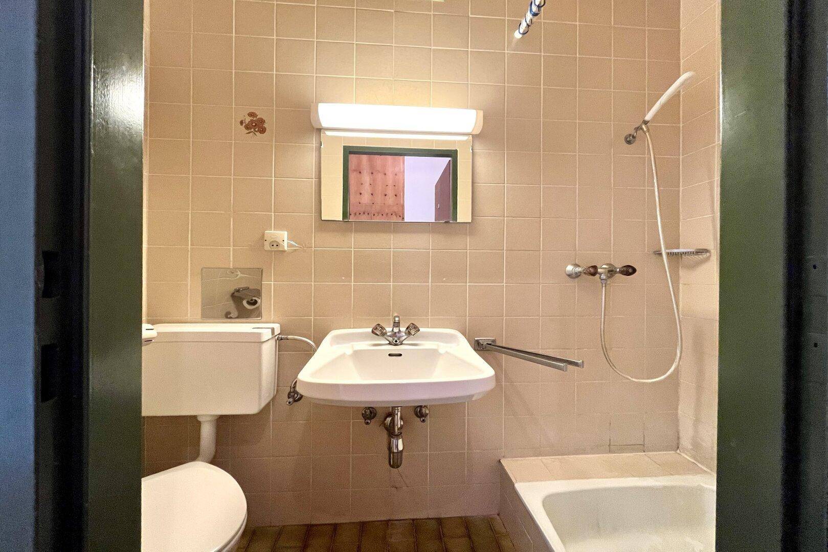 Badezimmer mit beigen Fliesen, Waschbecken, Toilette und einer Badewanne mit Duschfunktion.