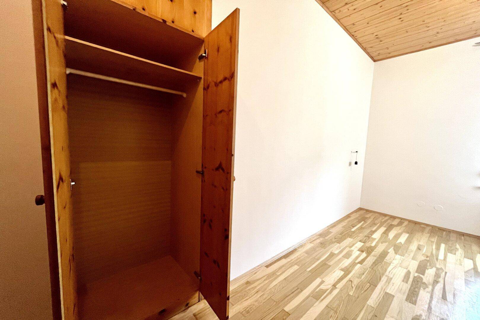 Leeres Zimmer mit Holzbalkendecke, Parkettboden und einem offenen Holzschrank.