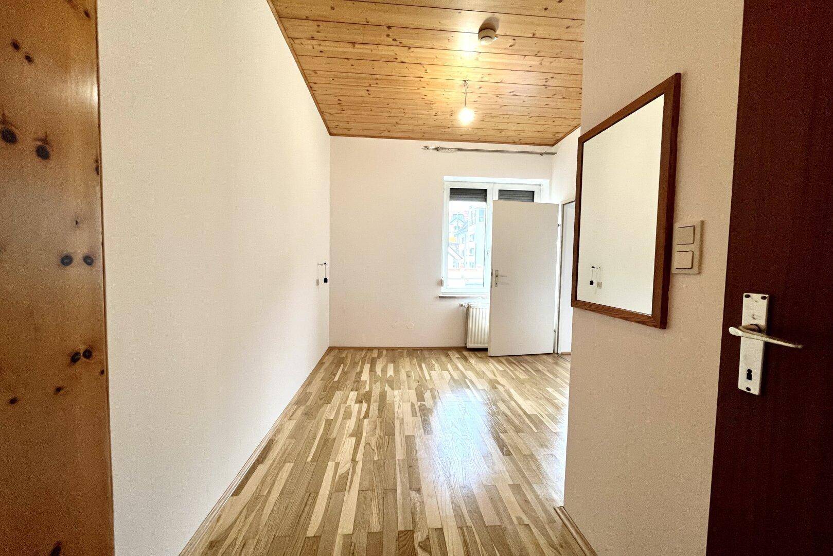 Heller Raum mit Holzbalkendecke, Parkettboden und einem Fenster, das viel Tageslicht hereinlässt.