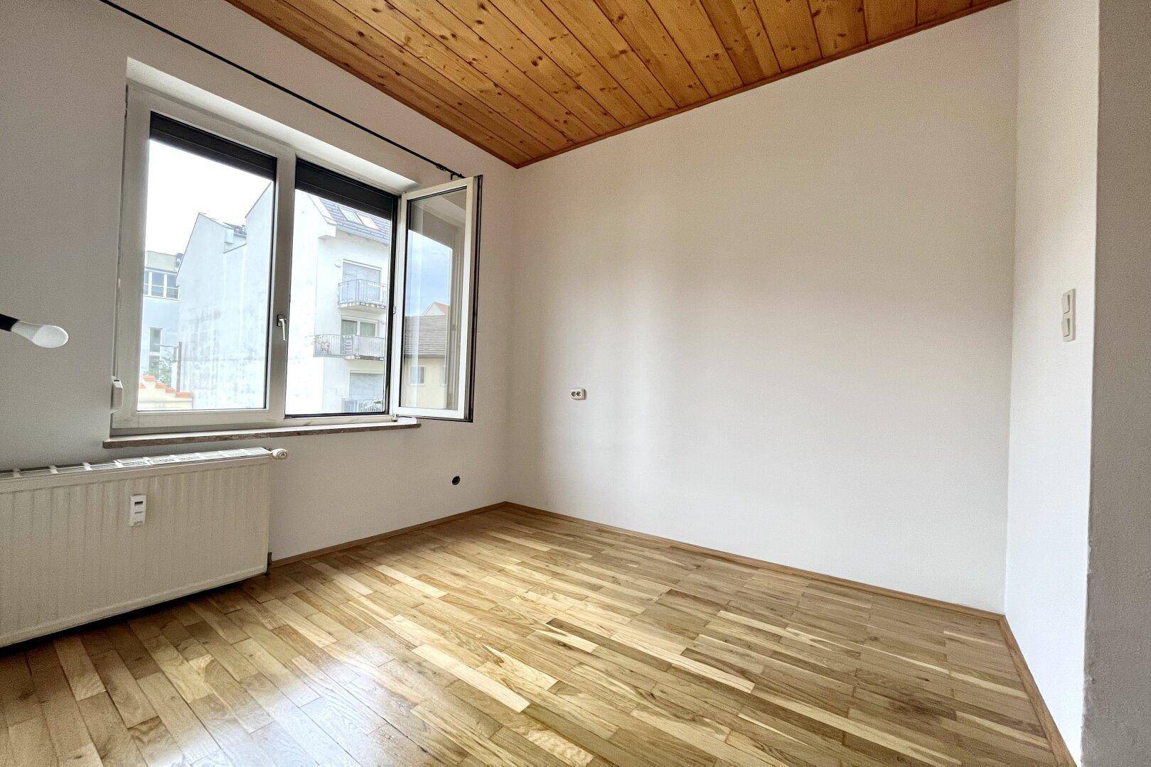 Leeres Zimmer mit Holzbalkendecke, Parkettboden und einem geöffneten Fenster, das frische Luft hereinlässt.