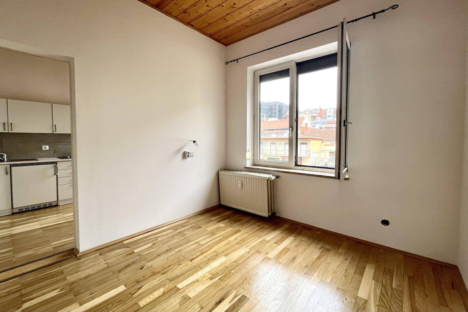 Heller Raum mit Holzbalkendecke und Parkettboden, der in den Küchenbereich übergeht.