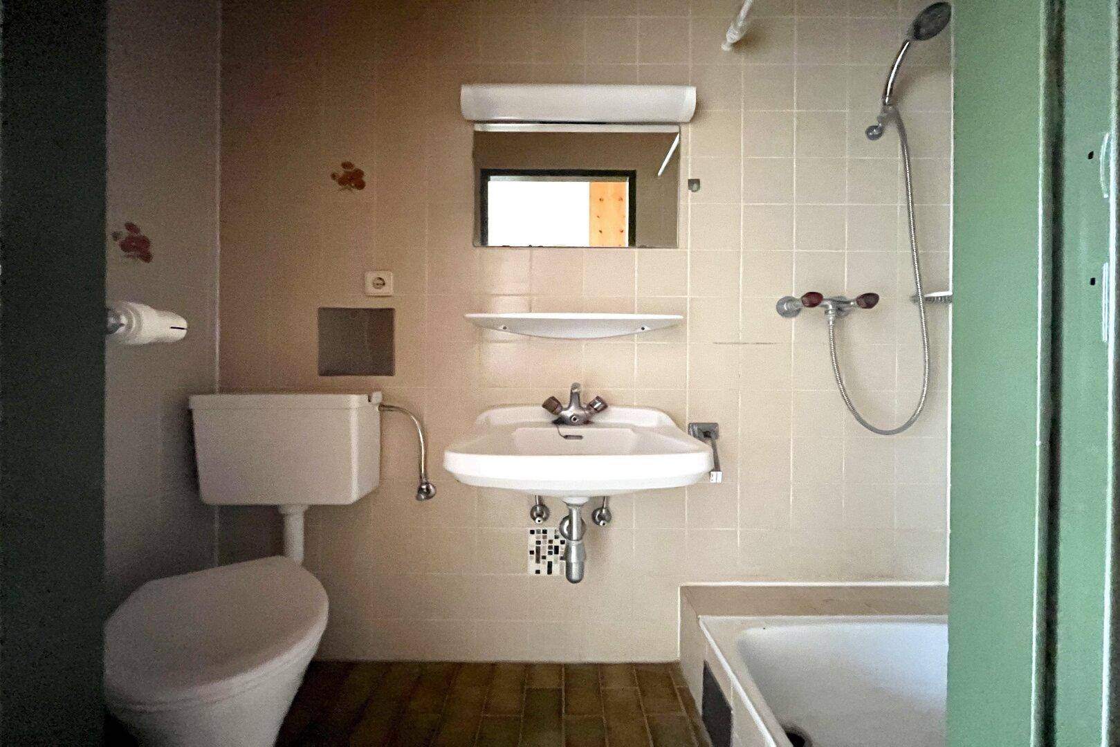 Badezimmer mit beigen Fliesen, Waschbecken, Toilette und einer Badewanne mit Duschfunktion.