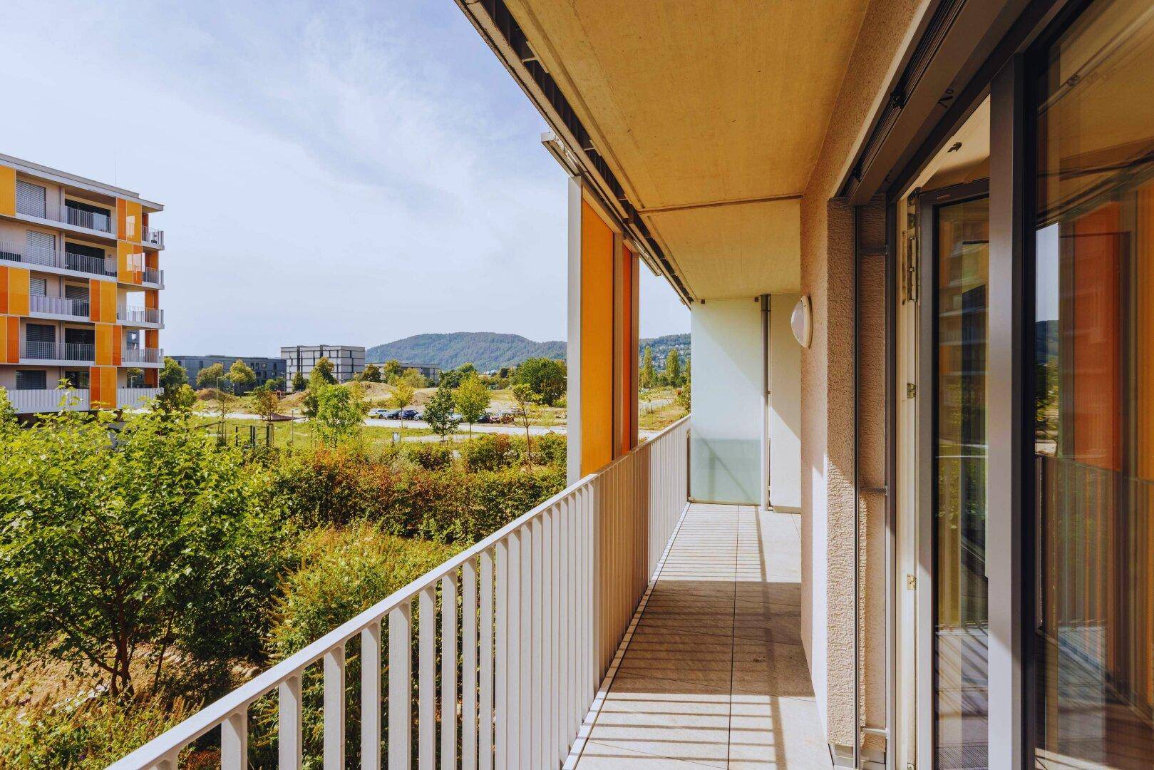 Langer Balkon mit Holzboden und Metallgeländer, bietet Ausblick auf Grünflächen und weitere Gebäude.