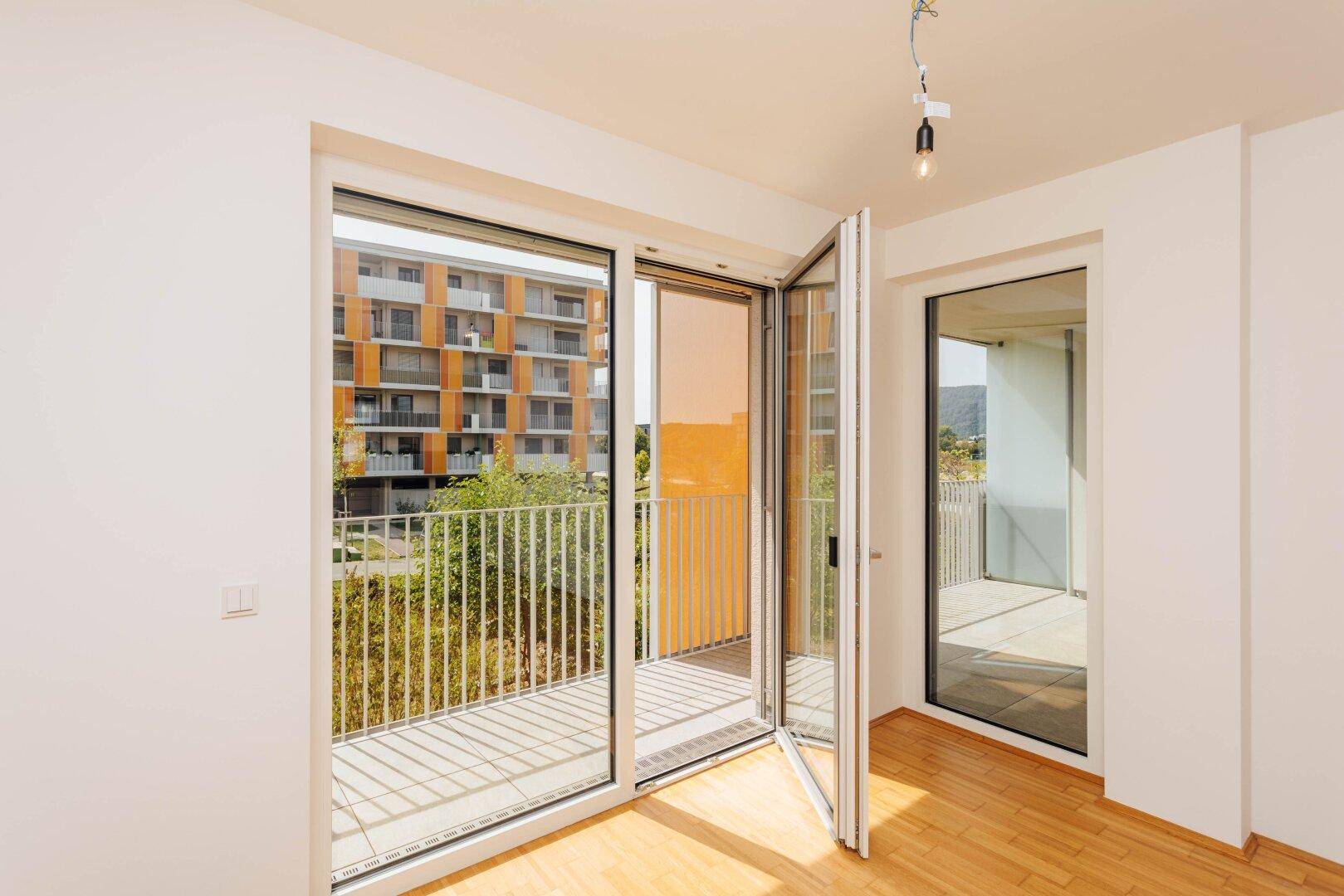 Heller Innenraum mit Holzboden und großen Fenstertüren, die zum Balkon führen und viel Licht hereinlassen.