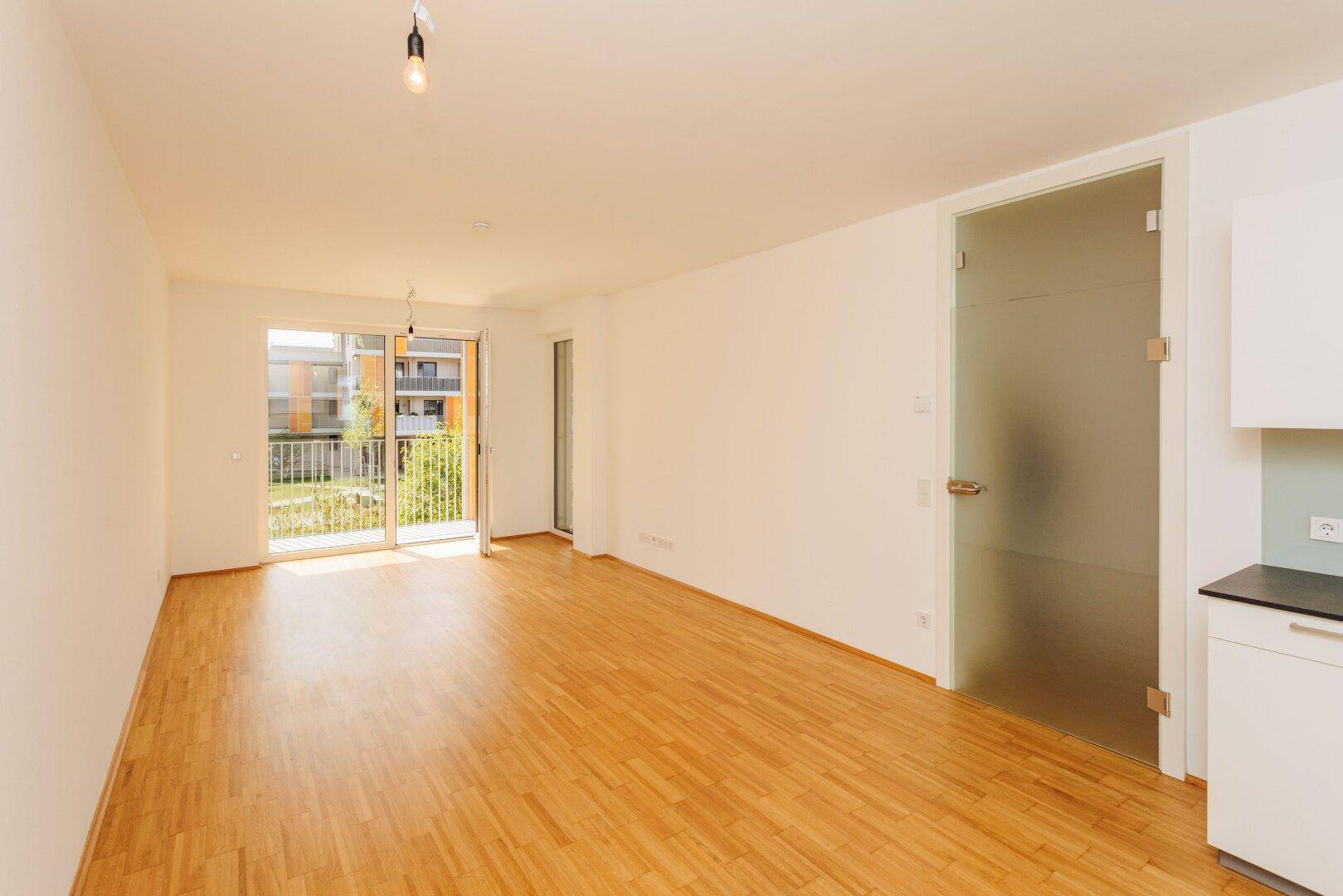 Leerer Wohnraum mit Holzboden, weißen Wänden und einer Glastür, die zu einem sonnigen Balkon führt.