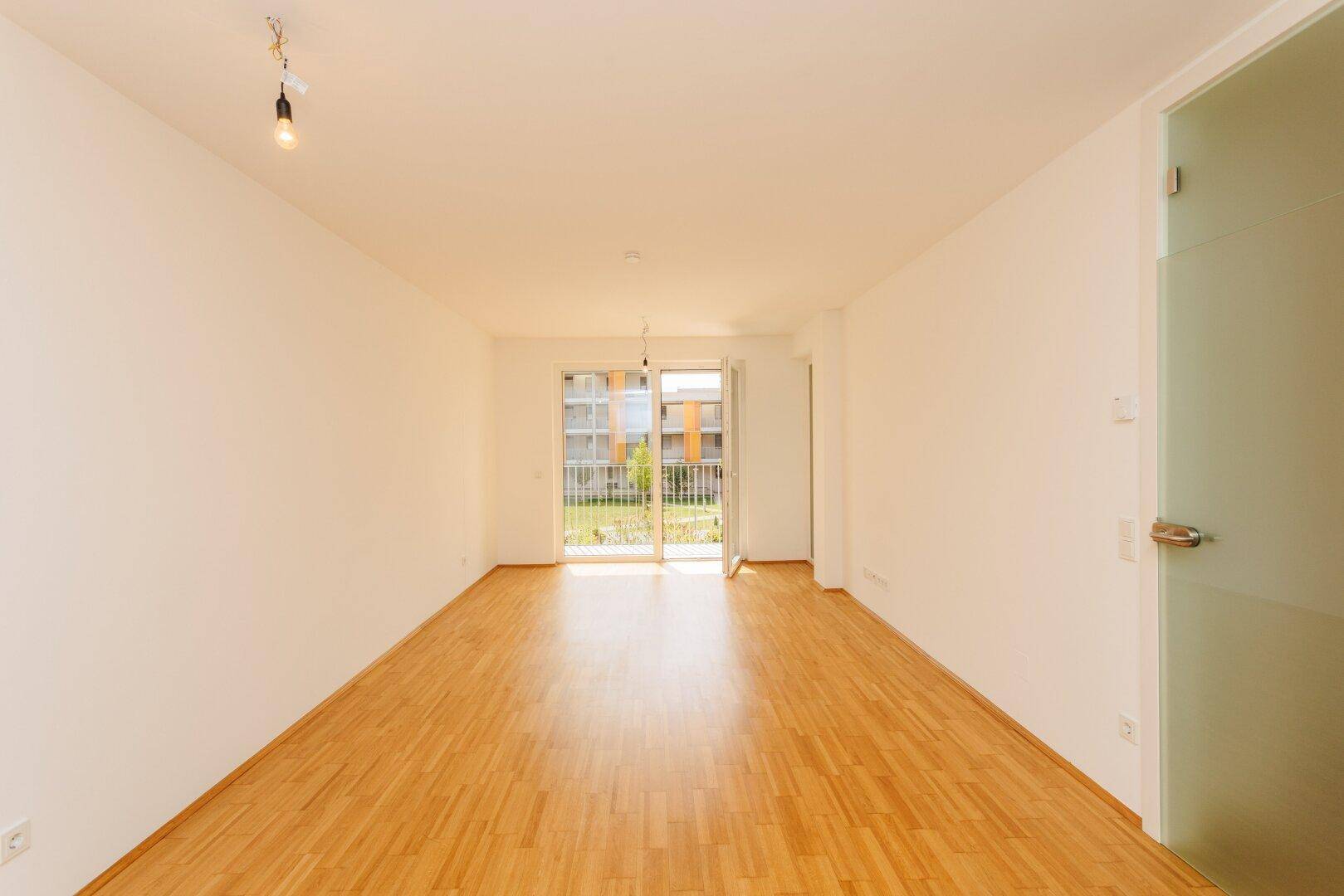 Heller, leerer Wohnraum mit Holzboden und einer Glastür, die zu einem sonnigen Außenbereich führt.