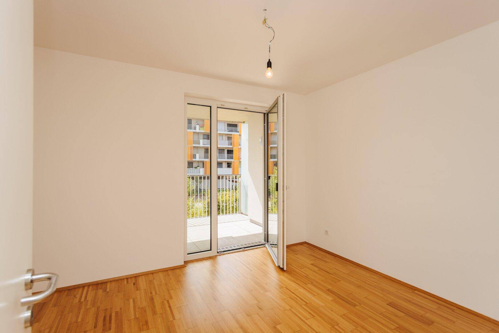 Leeres Schlafzimmer mit Holzboden und großen Fenstertüren, die zum Balkon führen und viel Licht spenden.
