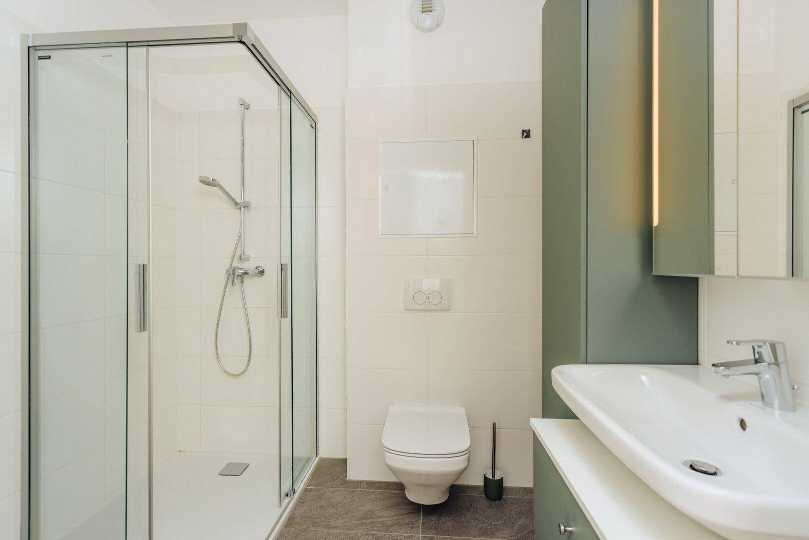 Modernes Badezimmer mit begehbarer Dusche, wandhängendem WC und grünem Waschtischunterschrank für Stauraum.