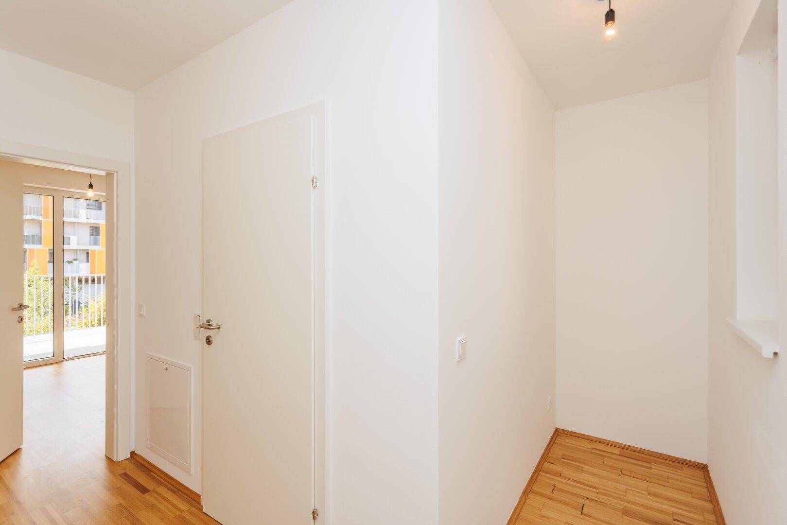 Heller Eingangsbereich mit Holzboden und weißen Wänden, der zu weiteren Räumen der Wohnung führt.