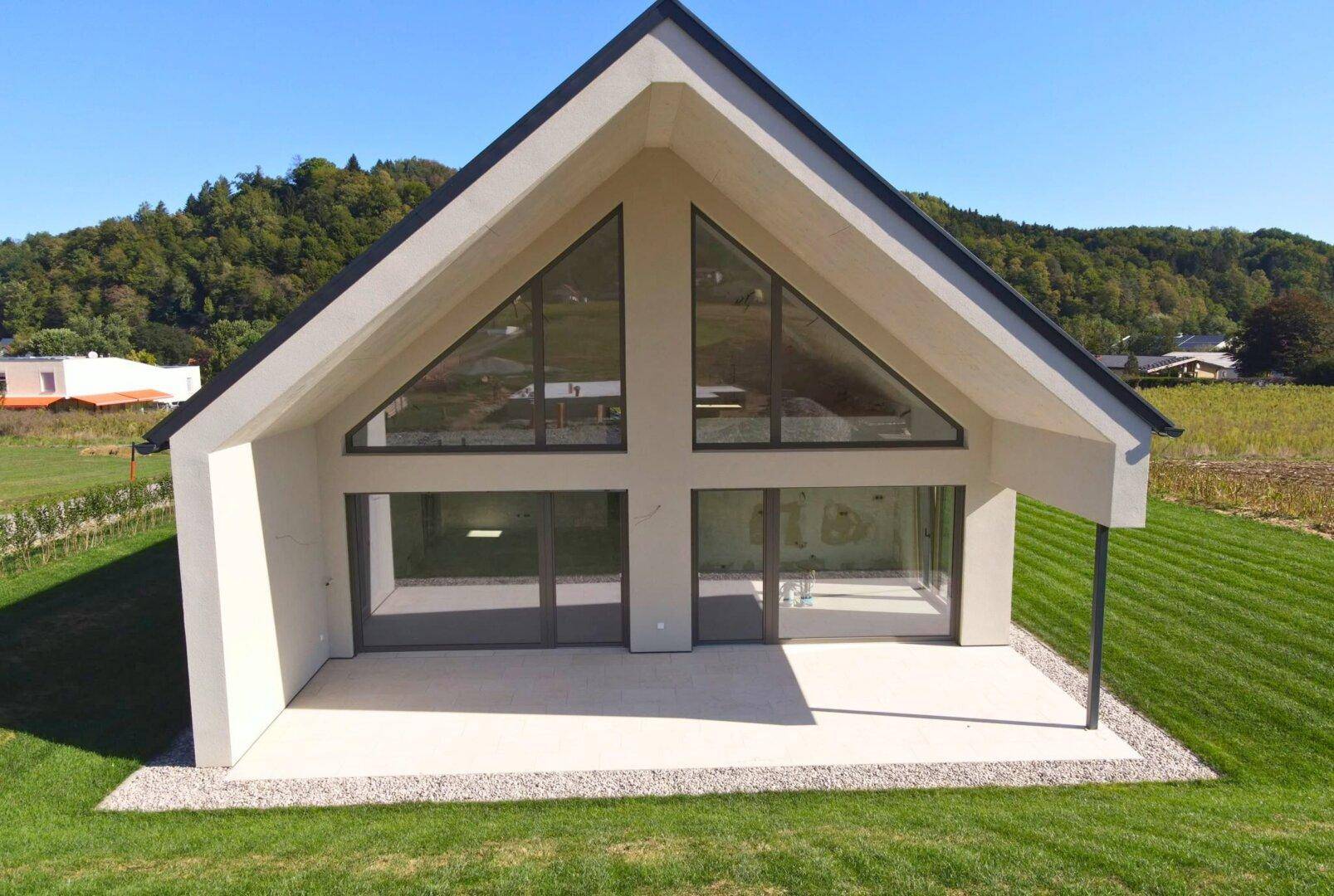 Frontansicht des modernen Hauses mit markanten Giebelfenstern und einer großzügigen, sonnigen Terrasse.