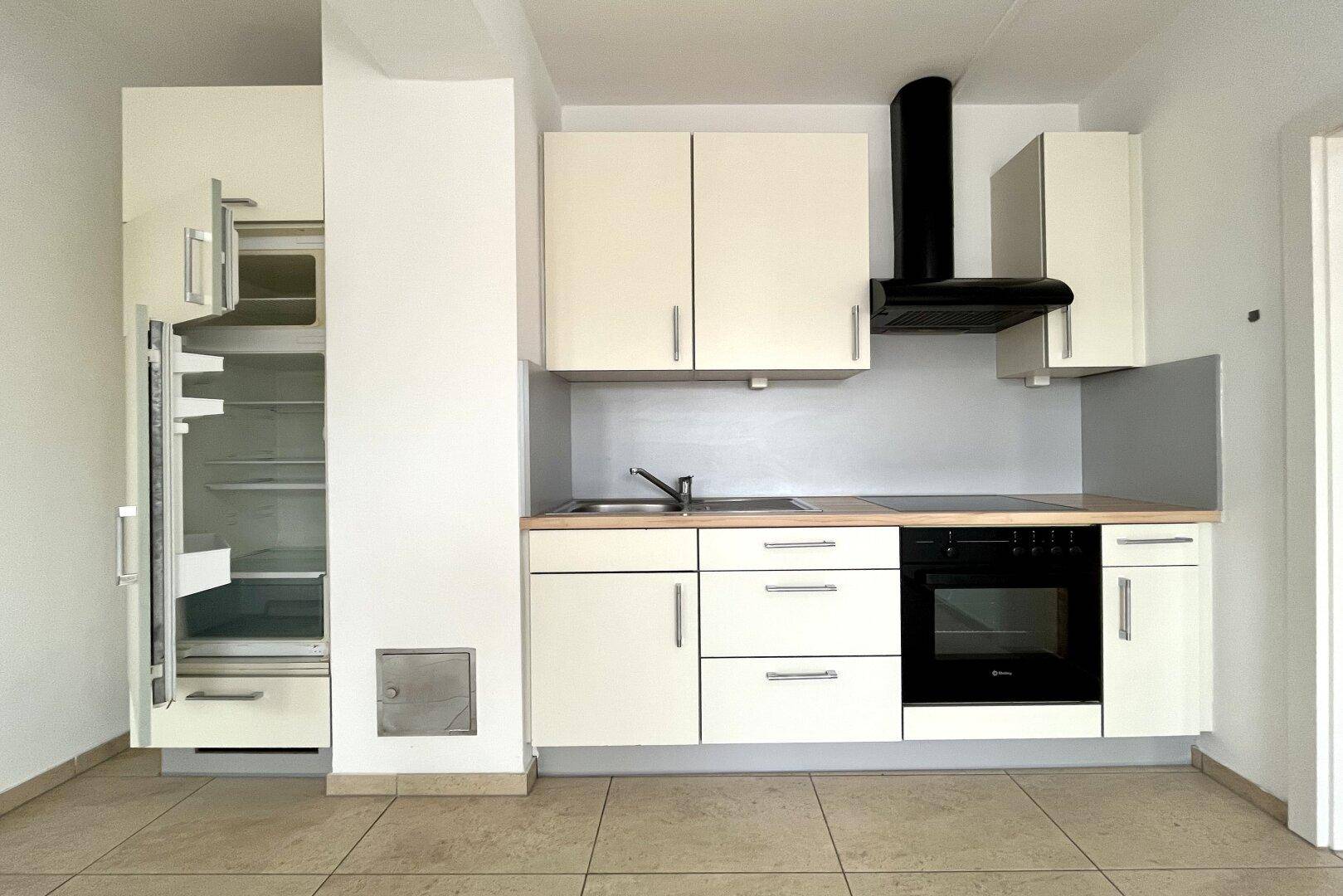 Kleine Küchenzeile mit weißen Schränken, Spüle, Herd und Kühlschrank in einem Wohnraum.