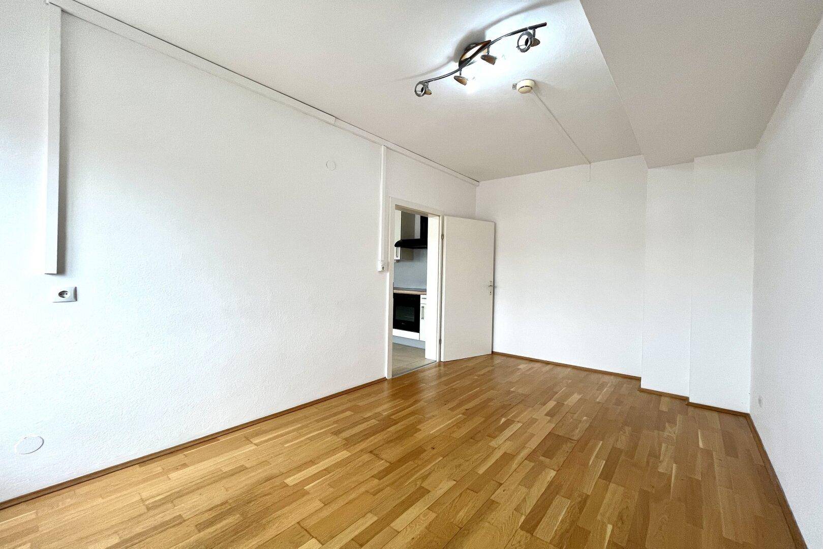 Heller Raum mit Holzparkettboden und Zugang zu einem weiteren Zimmer mit Küchenzeile.