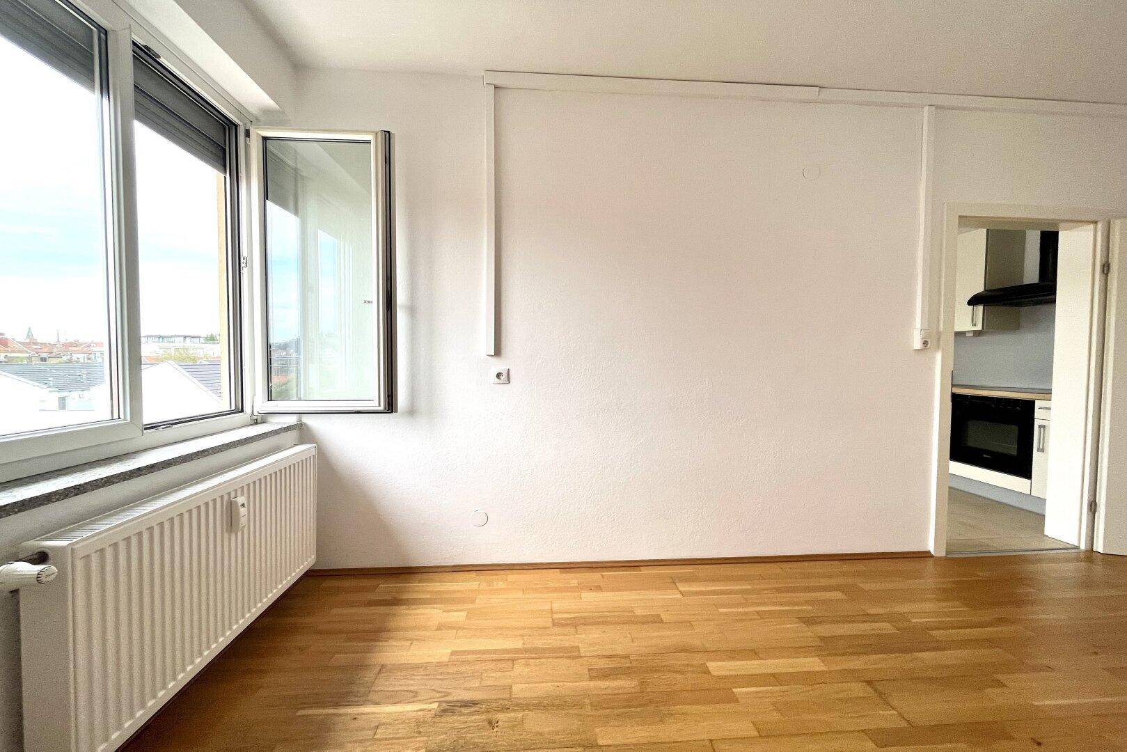 Geräumiger Raum mit Holzparkettboden, Heizkörper und großem Fenster mit Rollladen.