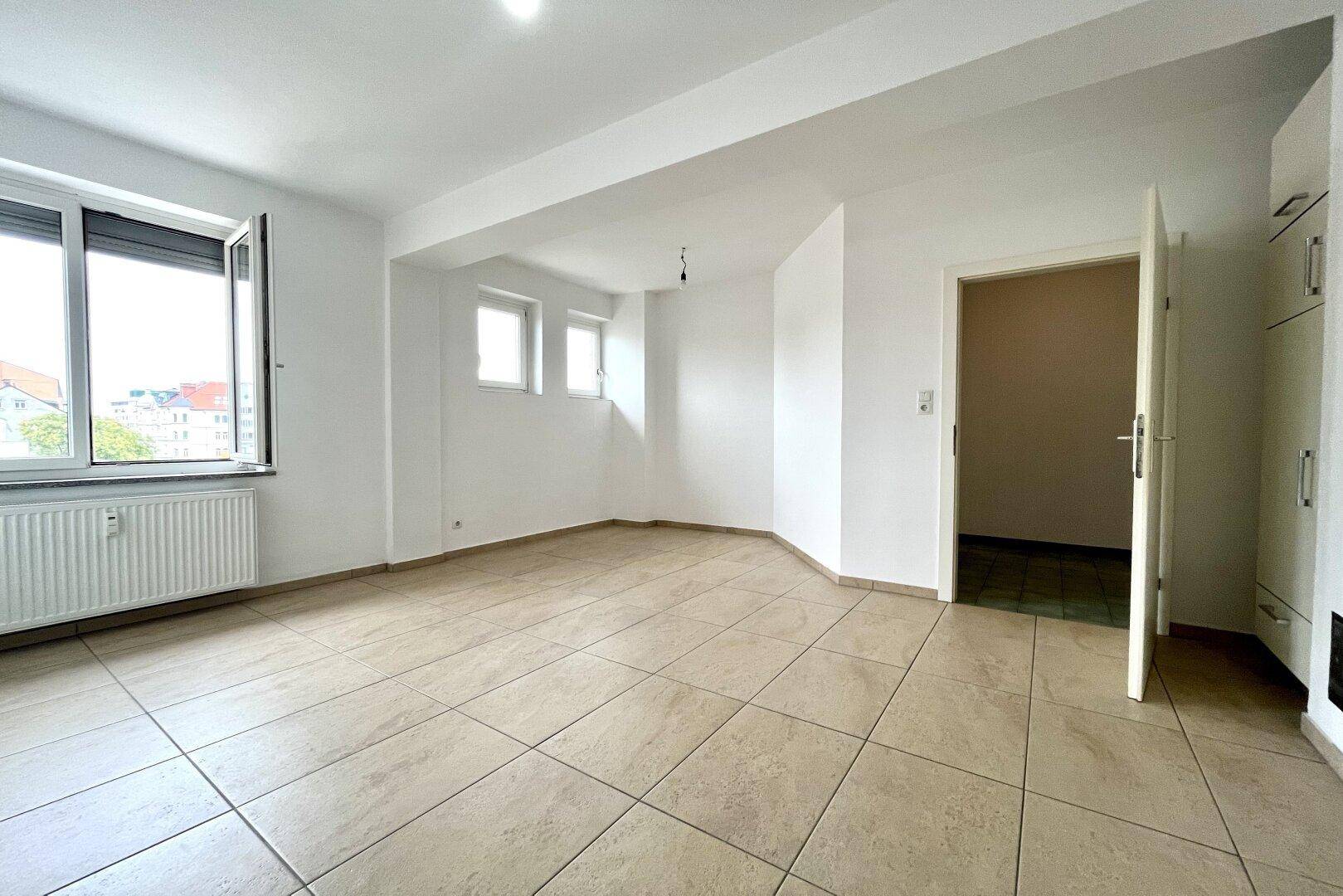 Großer, heller Raum mit Fliesenboden und zwei Fenstern, ideal als Wohn-Essbereich.