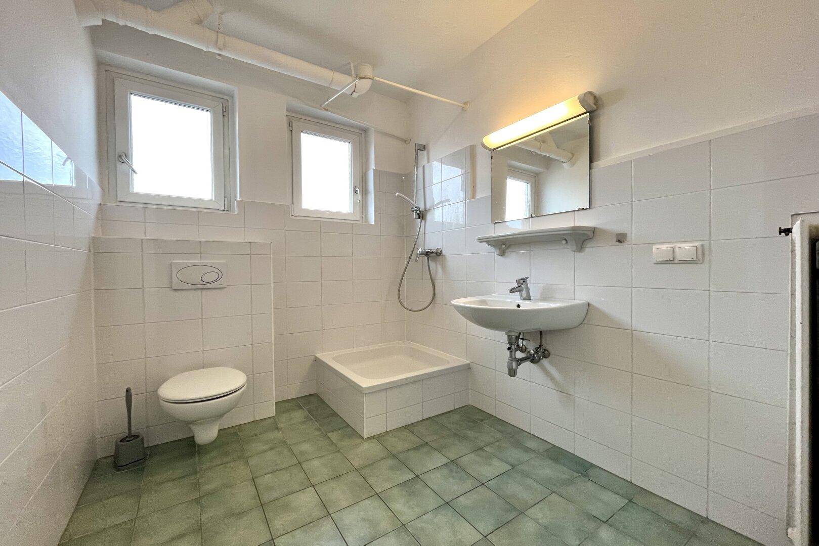 Badezimmer mit grünen Fliesen, Dusche, WC, Waschbecken und zwei Fenstern.