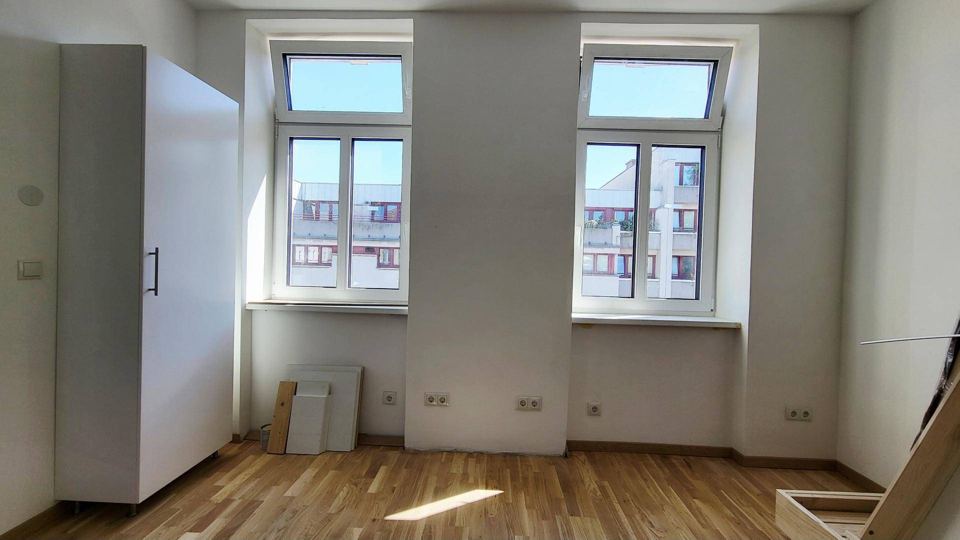 Unmöblierter Raum mit Holzfußboden, weißen Wänden und zwei großen Fenstern.
