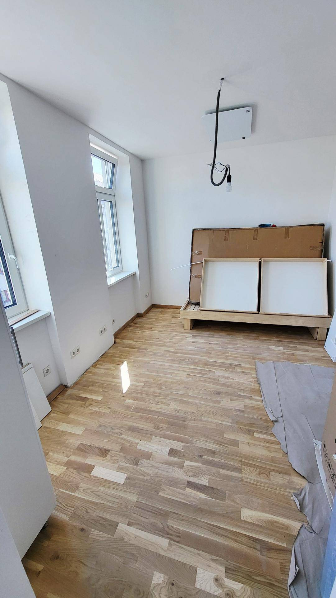 Heller Raum mit Holzfußboden und zwei Fenstern, derzeit unmöbliert mit Baumaterialien.
