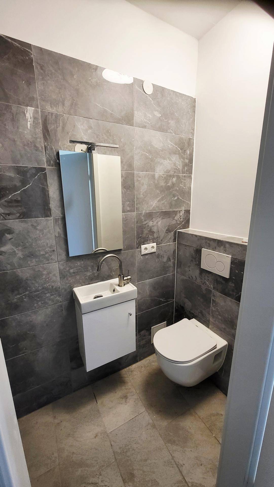 Modernes Gäste-WC mit grauen Fliesen, wandhängendem WC und kleinem Waschbecken mit Spiegelschrank.