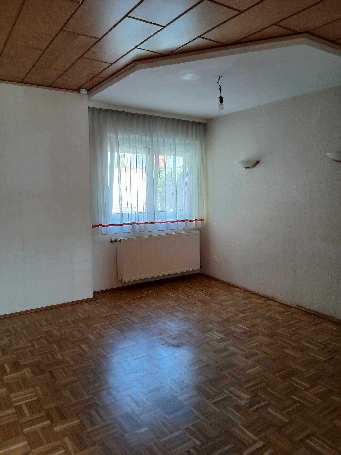 Geräumiges Zimmer mit Parkettboden, Holzdecke und Fenster mit Heizkörper.