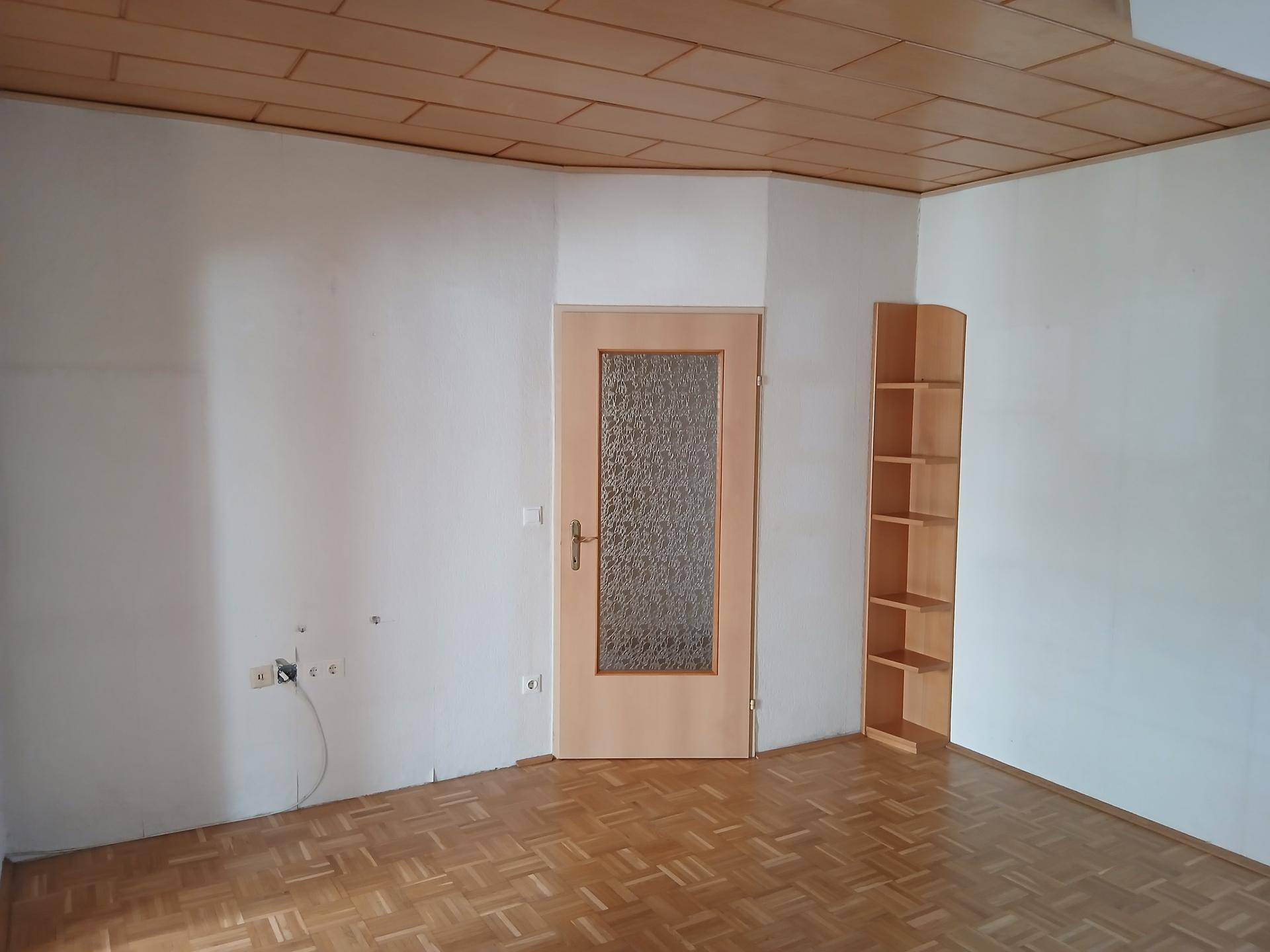 Leeres Zimmer mit Parkettboden, Holzdecke und einer Tür mit Glaseinsatz.