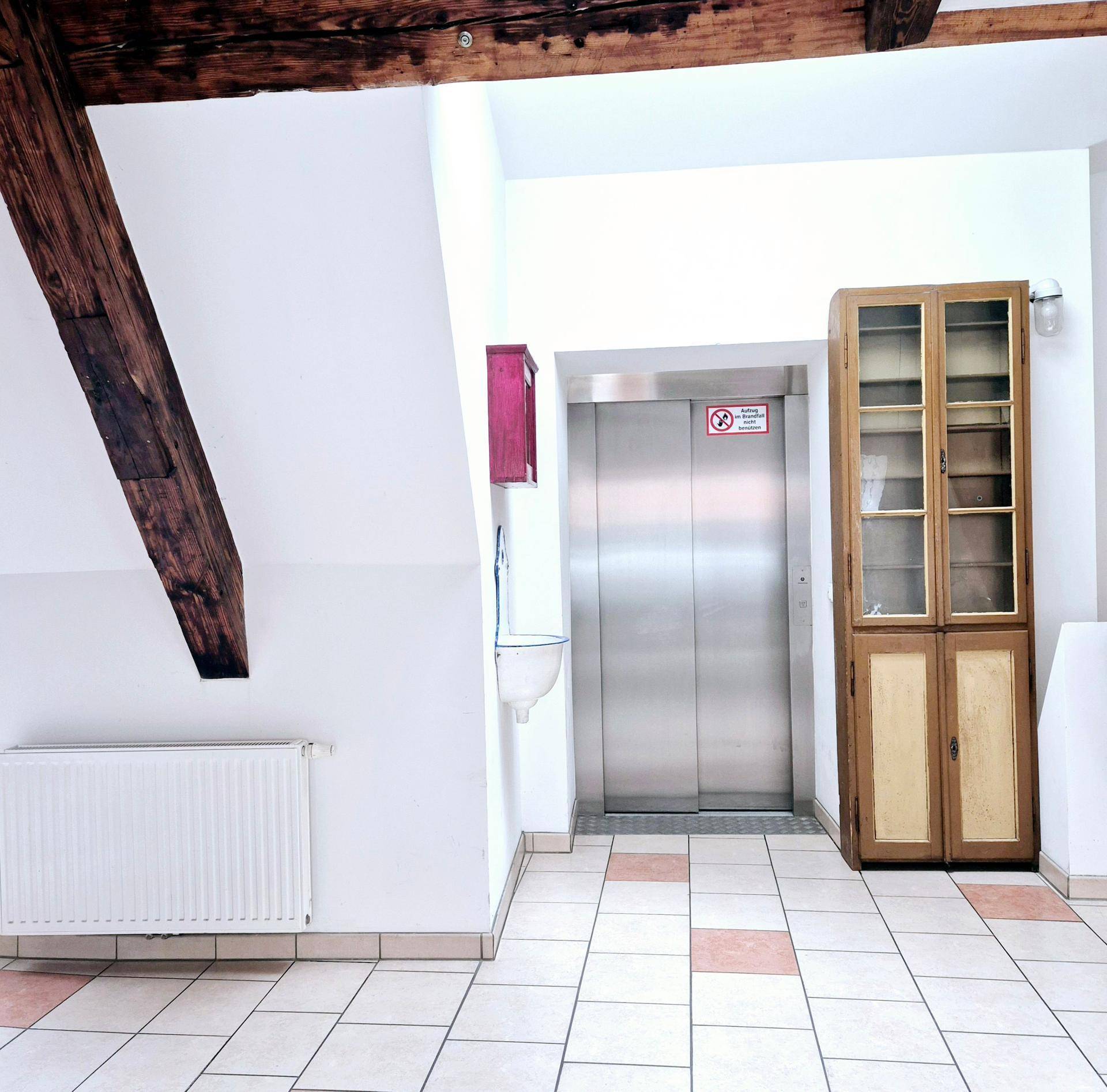 Gemeinschaftsbereich mit Aufzug, Fliesenboden und sichtbaren Holzbalken.