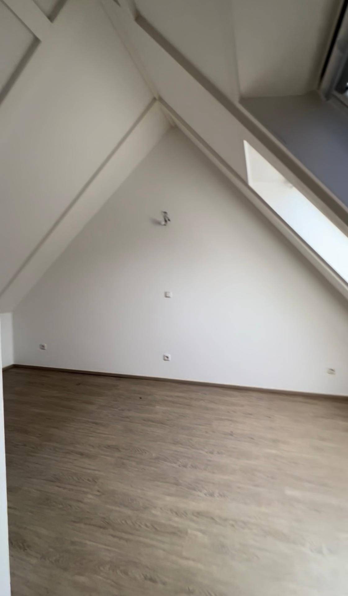 Leerer Raum im Dachgeschoss mit schrägen Wänden, hellem Boden und einem Dachfenster für natürliches Licht.