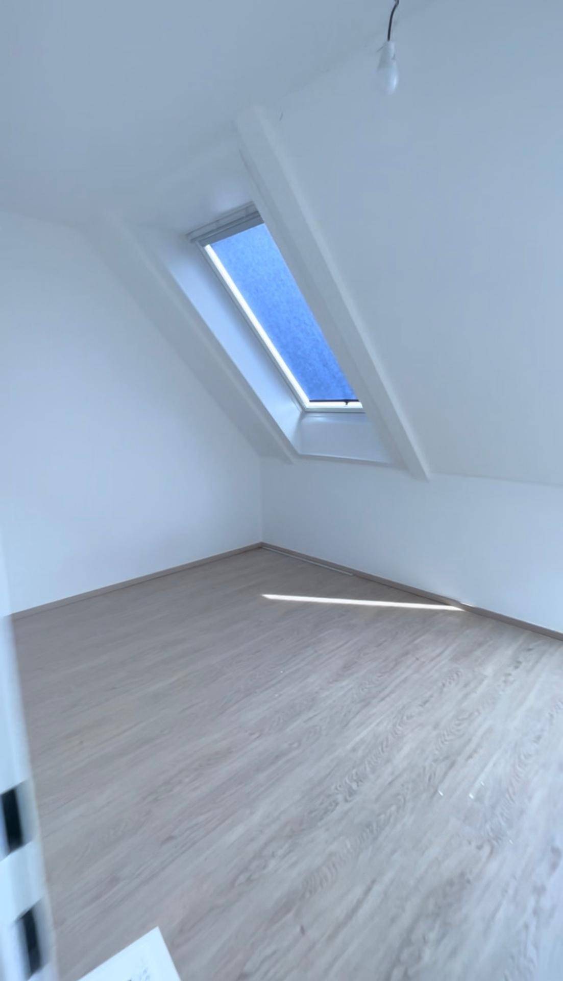 Heller Dachgeschossraum mit schrägen Wänden, Laminatboden und einem Dachfenster mit blauer Jalousie.