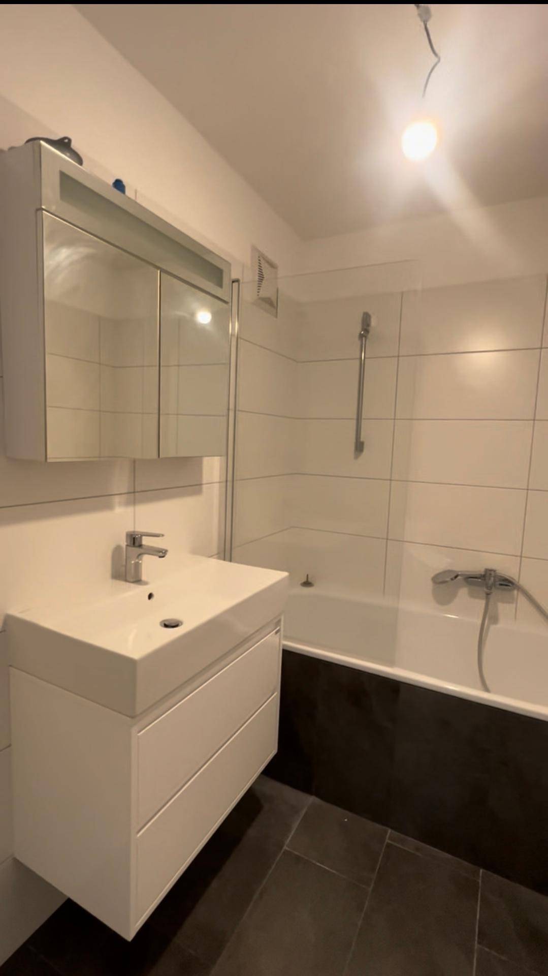 Helles Badezimmer mit Badewanne, Duschfunktion, modernem Waschtisch und Spiegelschrank auf dunklen Fliesen.