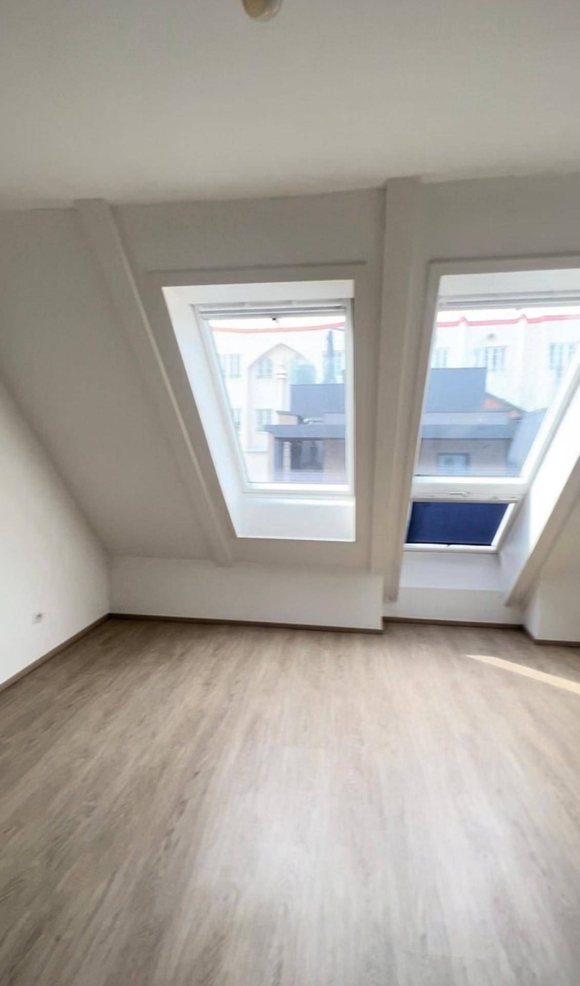 Heller Dachgeschossraum mit zwei Dachfenstern, die einen Blick auf die umliegenden Gebäude bieten.