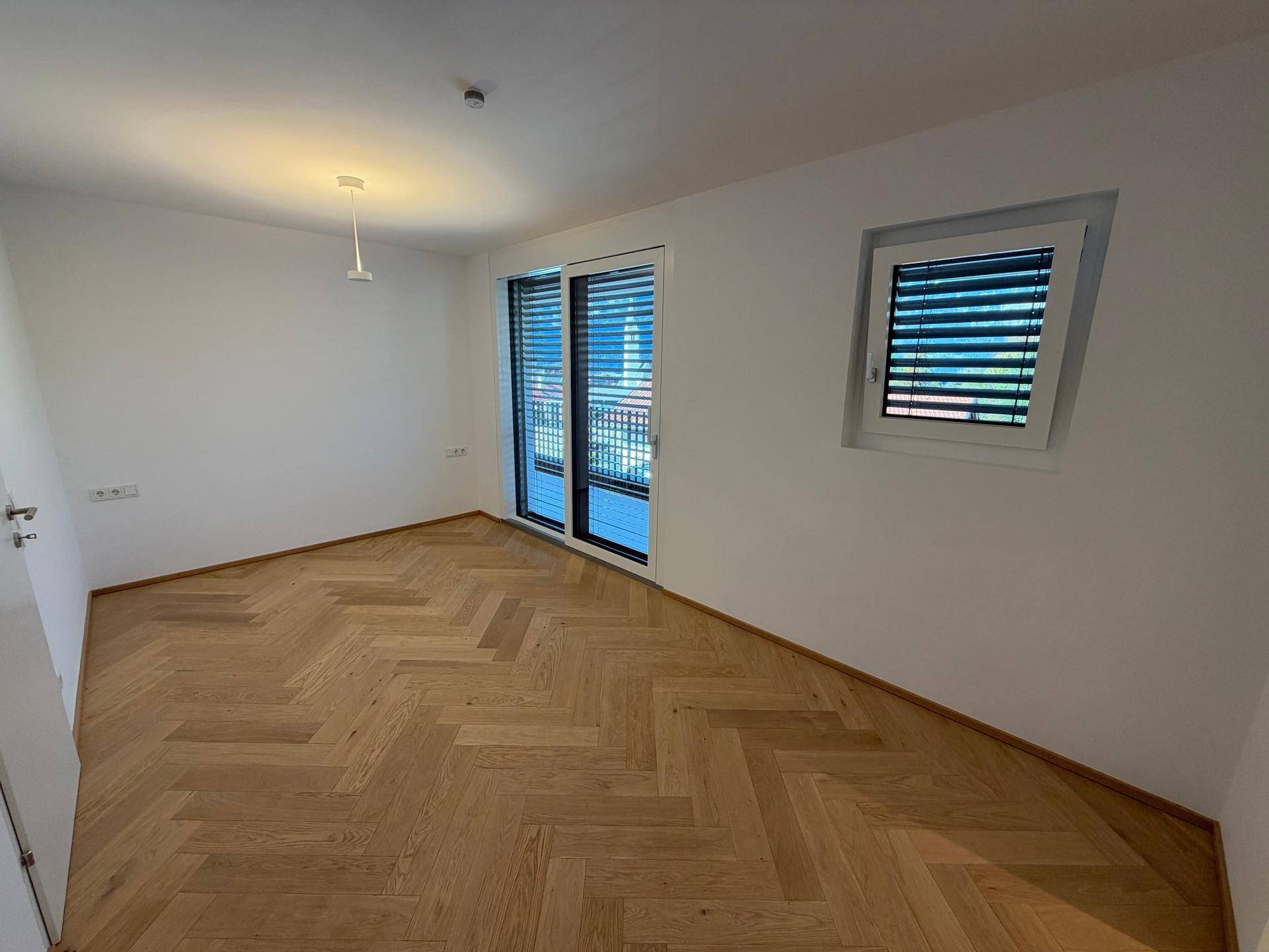 Geräumiges Schlafzimmer mit großen Fenstern, Fischgrätparkett und viel Tageslicht.