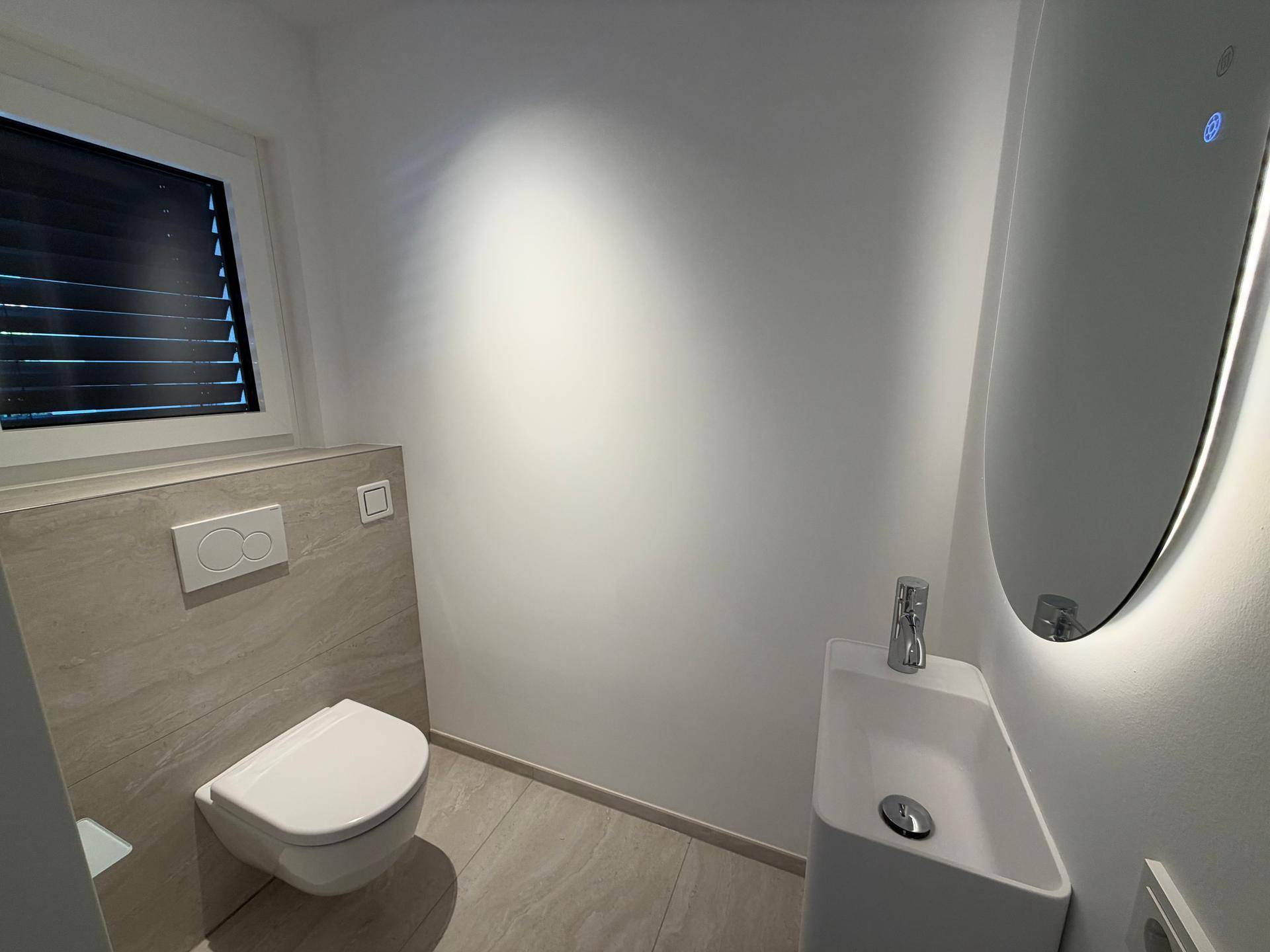 Modernes Gäste-WC mit Wand-WC, kleinem Waschbecken und beleuchtetem Spiegel.