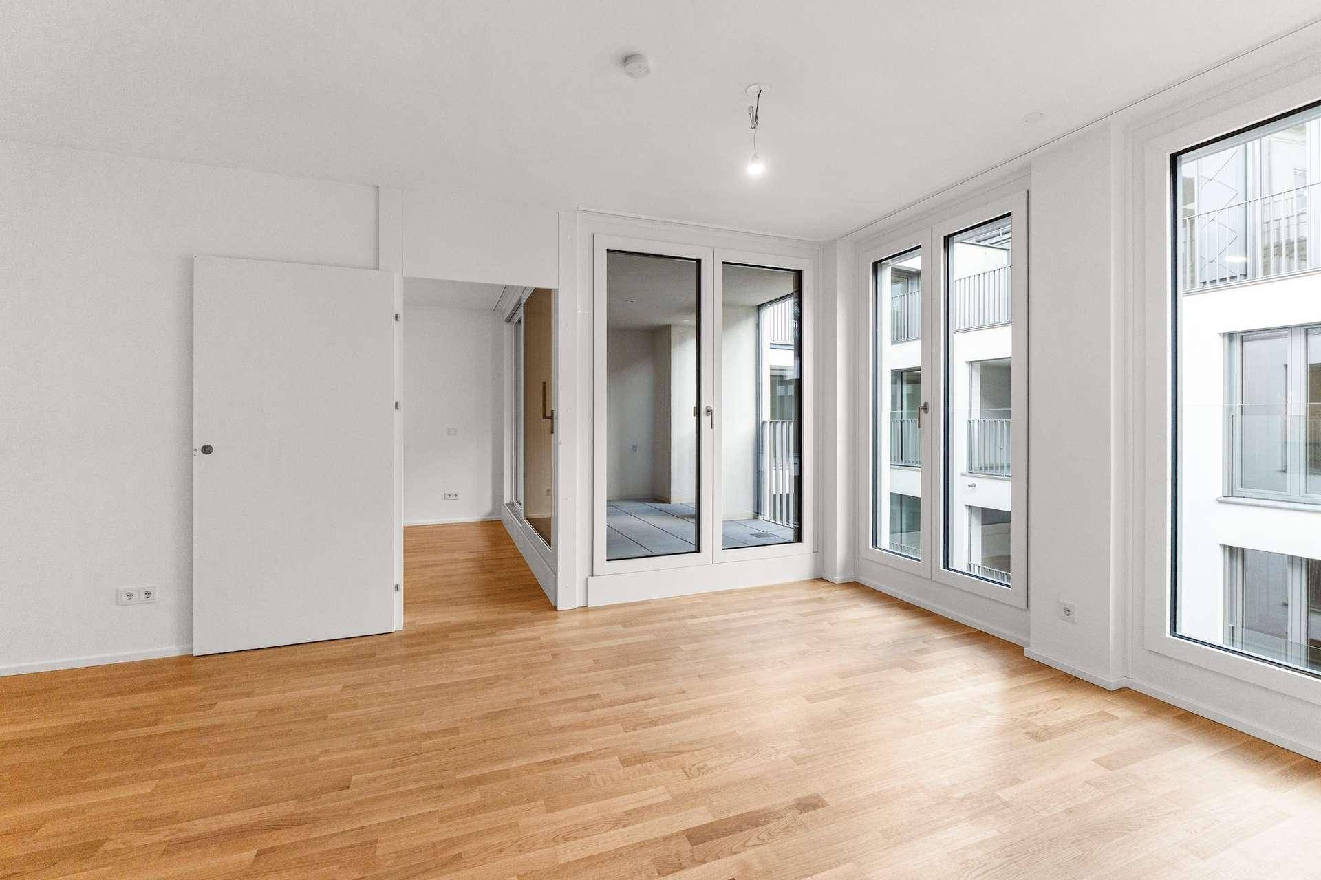 Helles, leeres Wohnzimmer mit Holzboden und großen Fenstern, die auf einen Innenhof blicken.