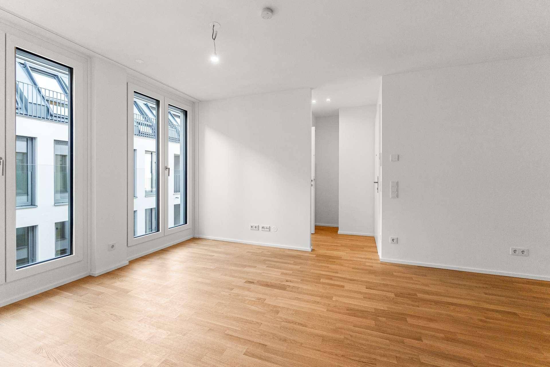 Heller, leerer Wohnraum mit Holzboden und großen Fenstern, die viel Tageslicht hereinlassen.