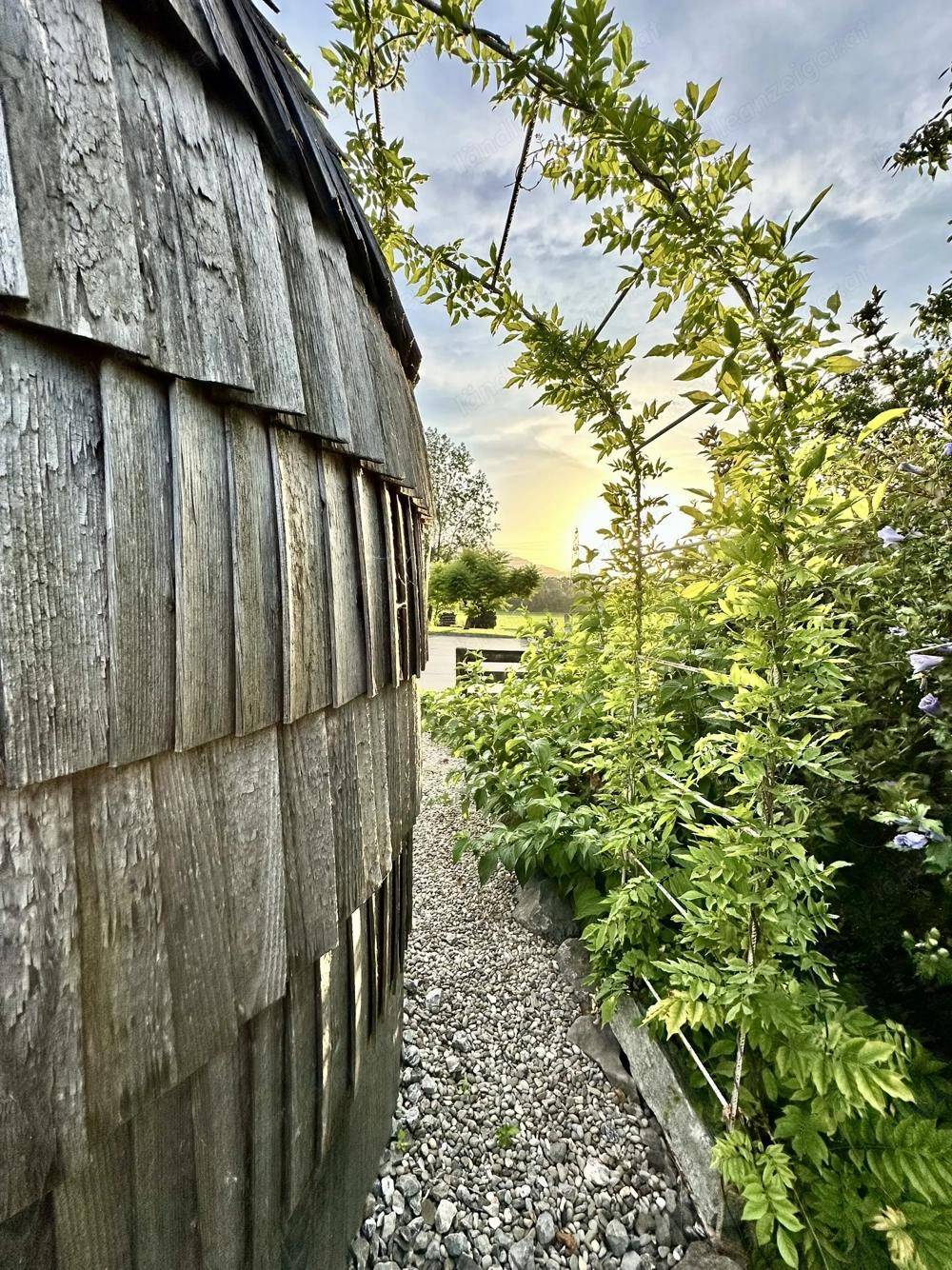 Seitenansicht des Gebäudes mit Holzschindeln und einem schmalen Kiesweg, umgeben von üppigem Grün im Sonnenuntergang.