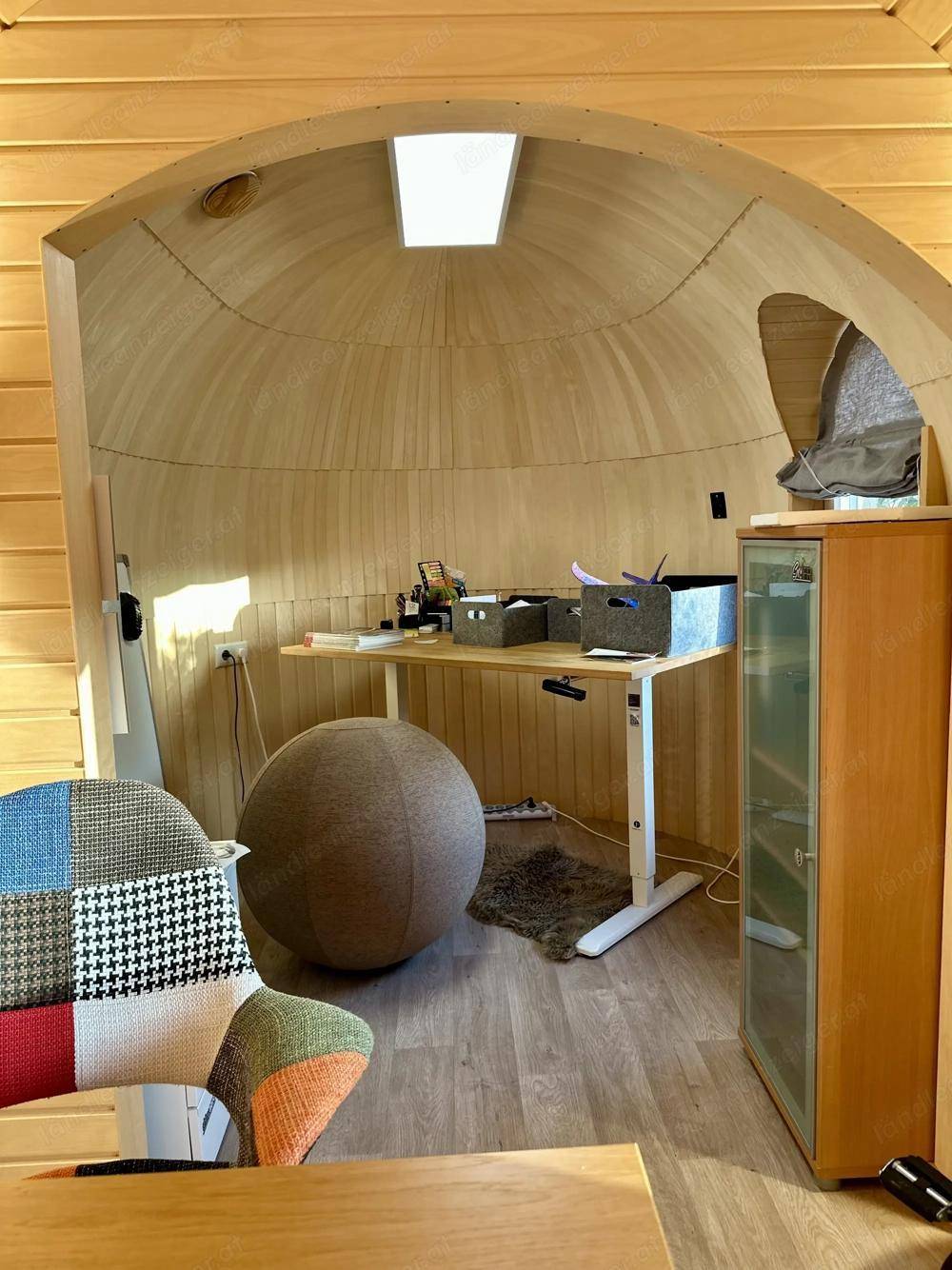Helles Büro mit gewölbter Holzdecke und -wänden, ausgestattet mit einem Schreibtisch und einem ergonomischen Ballstuhl.