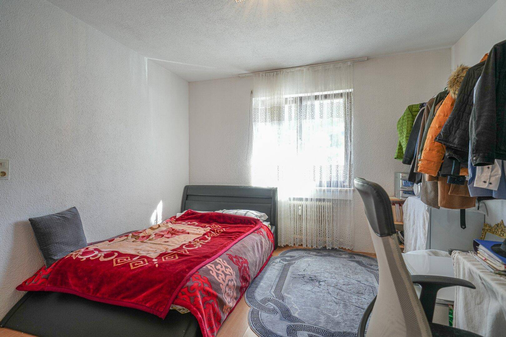 Schlafzimmer mit Einzelbett, Schreibtisch, Kleiderschrank und hellem Fenster, das viel Licht hereinlässt.