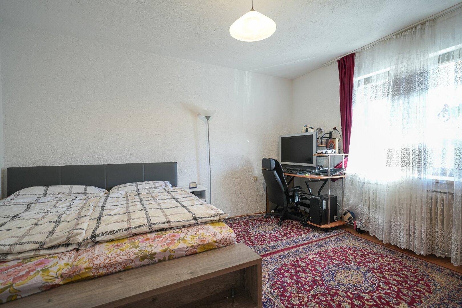 Schlafzimmer mit Doppelbett, Schreibtisch und Computer, großen Fenstern und zwei gemusterten Teppichen.