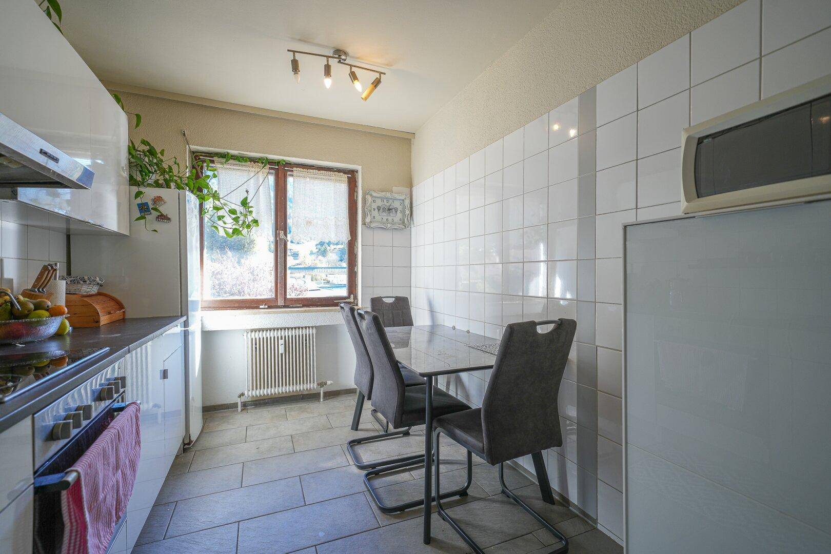 Heller Essbereich in der Küche mit Tisch und Stühlen, umgeben von weißen Fliesen und einem Fenster.