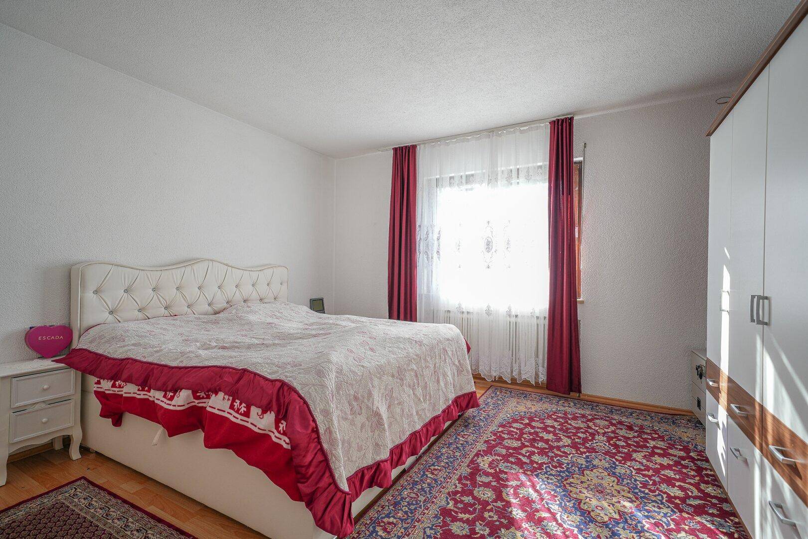 Geräumiges Schlafzimmer mit großem Doppelbett, weißem Kleiderschrank und einem Fenster mit roten Vorhängen.