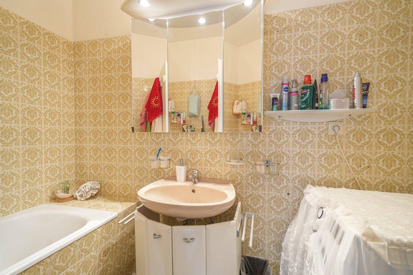 Badezimmer mit Badewanne, Waschbecken, Spiegelschrank und charakteristischen gelb-gemusterten Fliesen.
