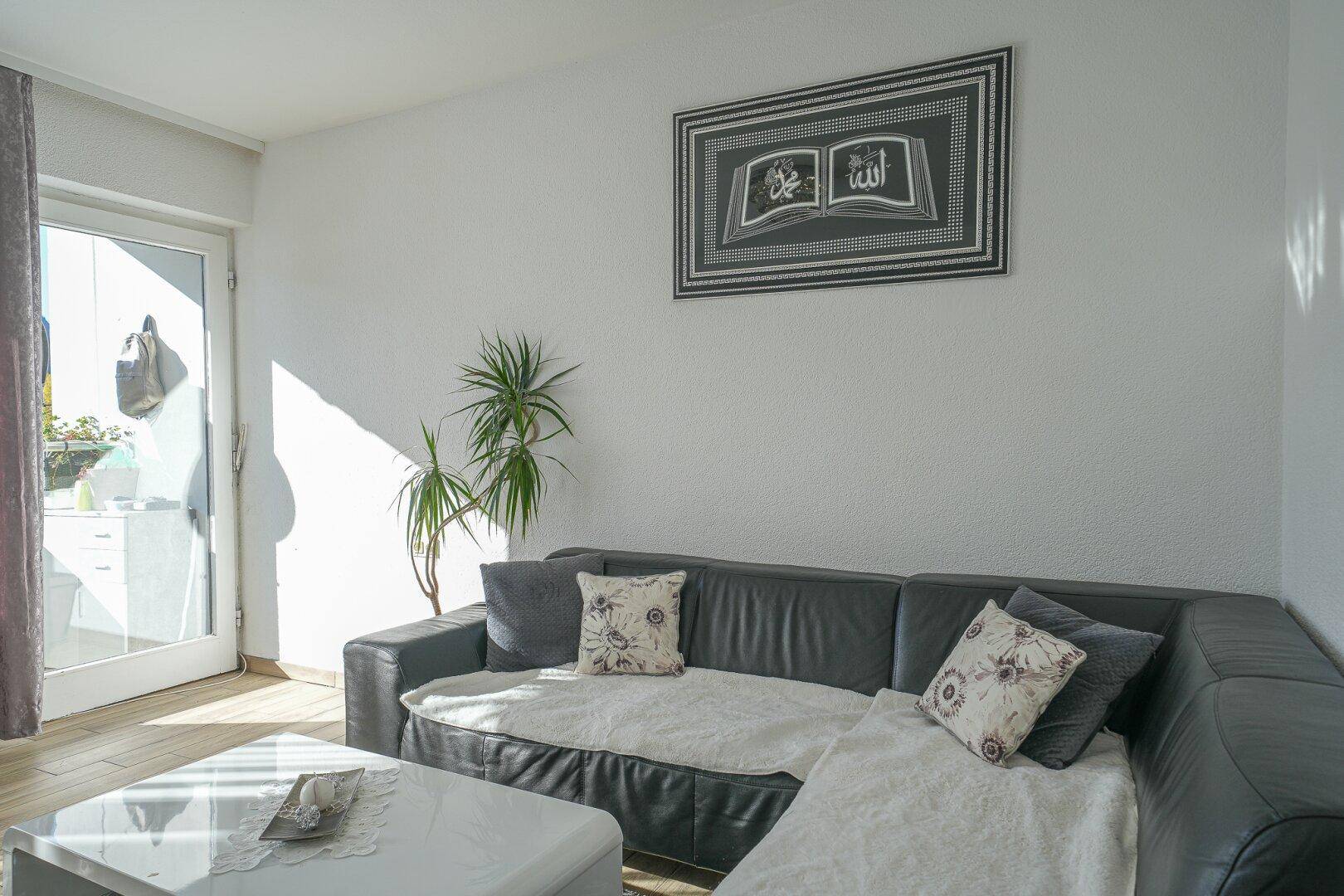 Helles Wohnzimmer mit schwarzem Ecksofa, weißem Couchtisch und Zugang zu einem Balkon oder einer Terrasse.