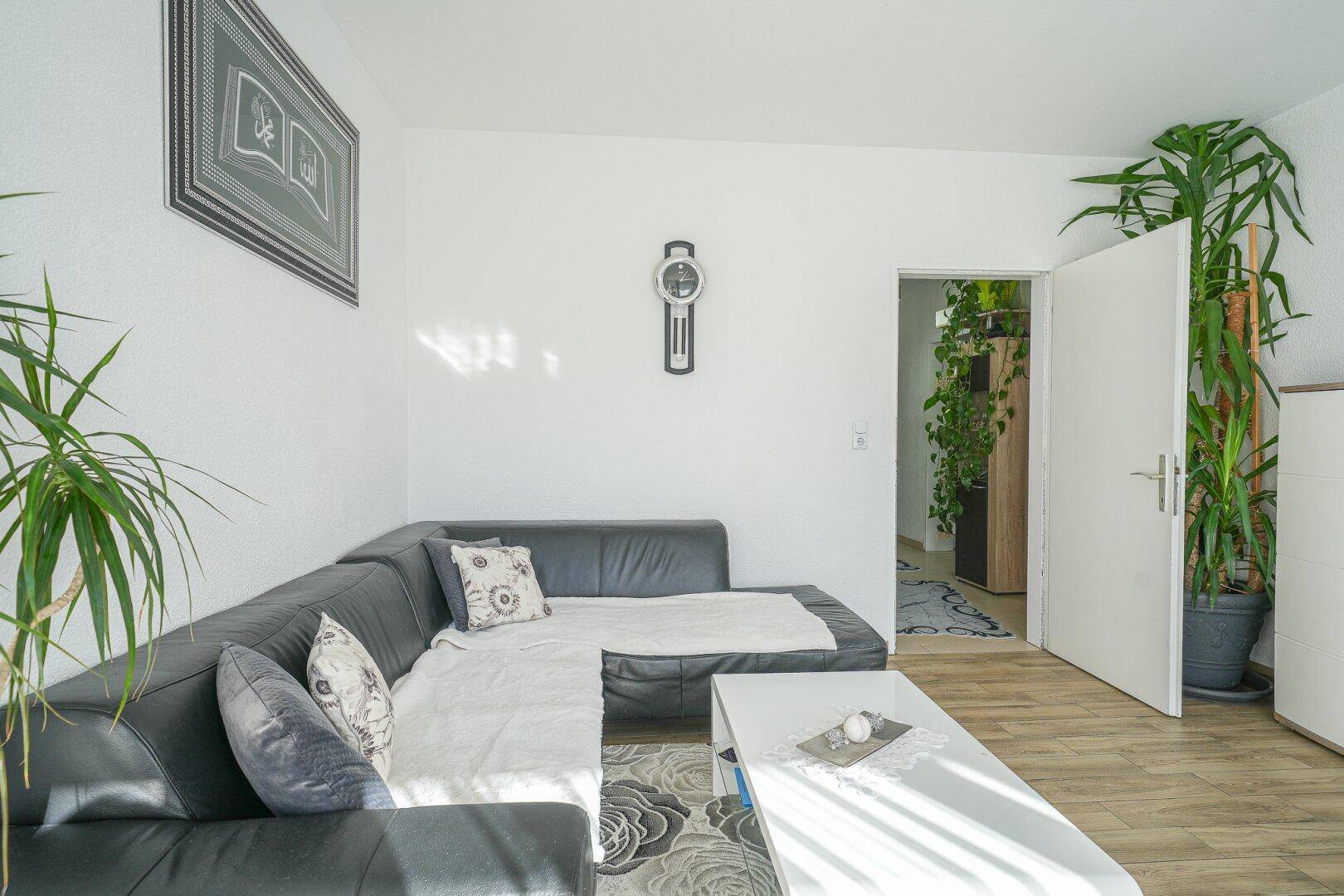 Gemütliches Wohnzimmer mit schwarzem Sofa, hellem Laminatboden und einem Durchgang zu einem weiteren Raum.
