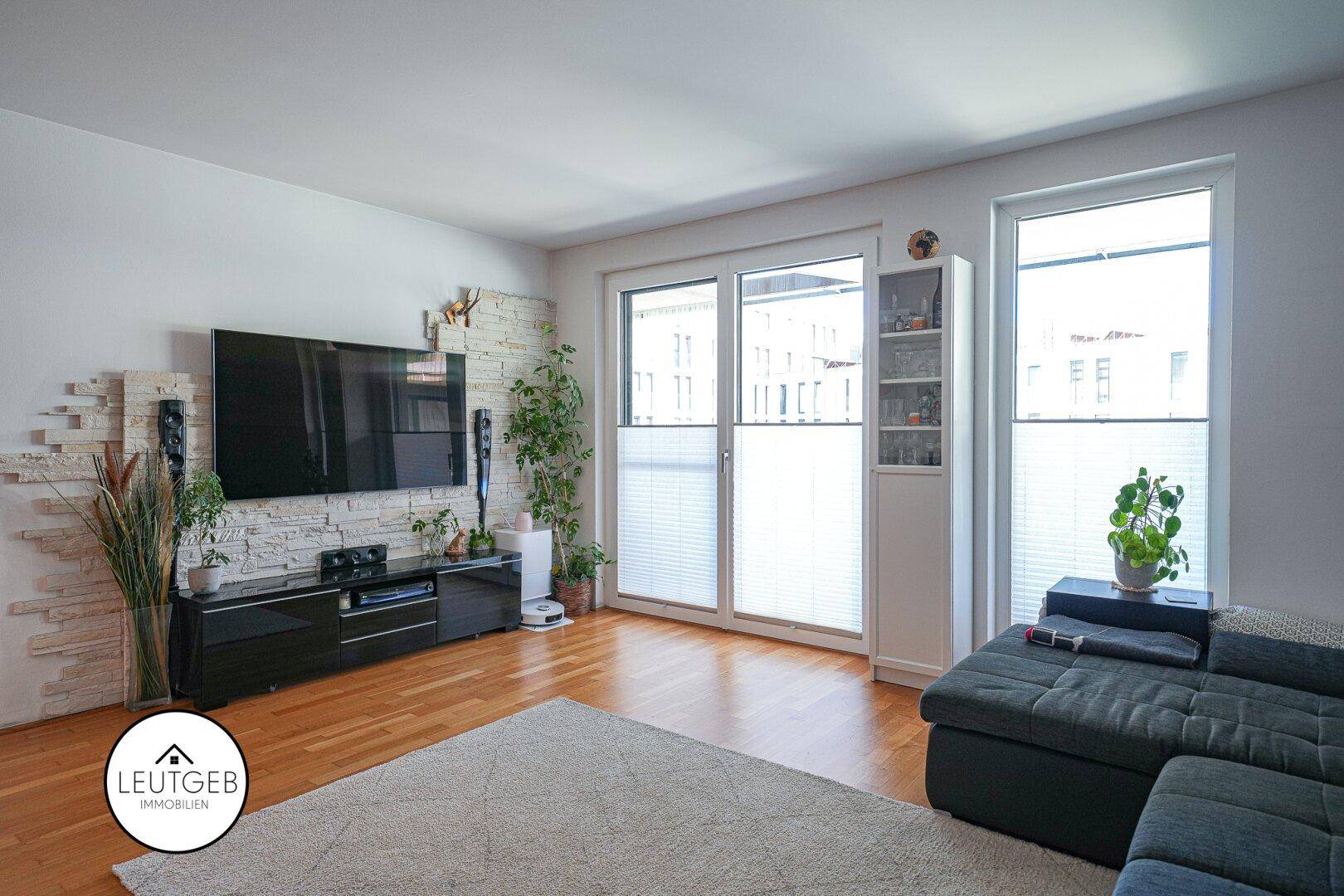 Gemütliches Wohnzimmer mit großem Fernseher, Steinwandakzent und Zugang zum Balkon.
