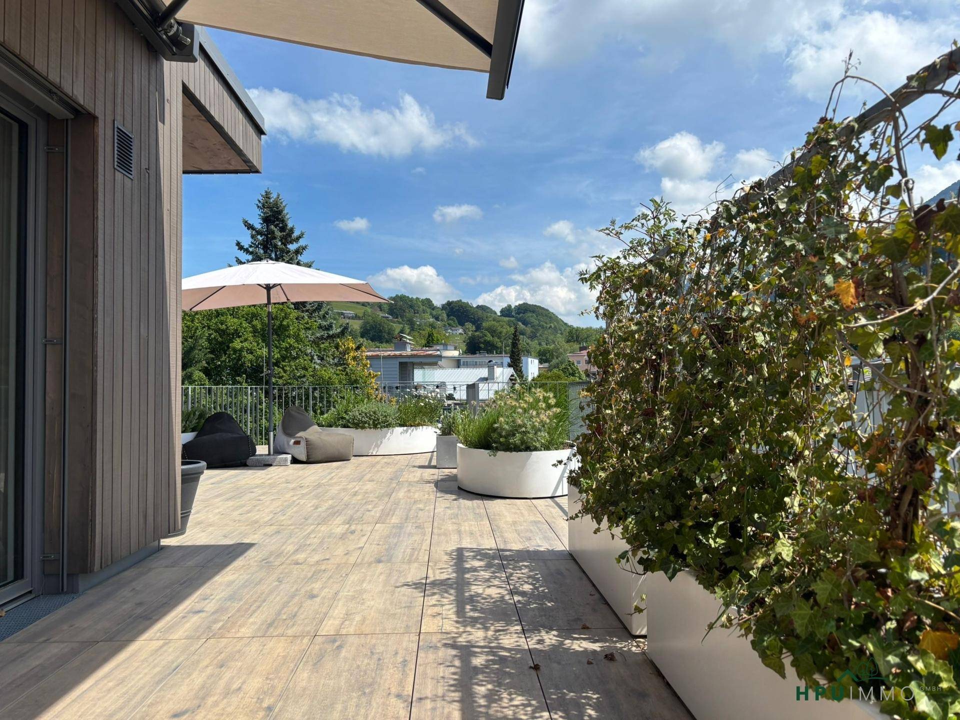 Weitläufige Dachterrasse mit Pflanzen, Sonnenschirm und Blick auf die grüne Umgebung.