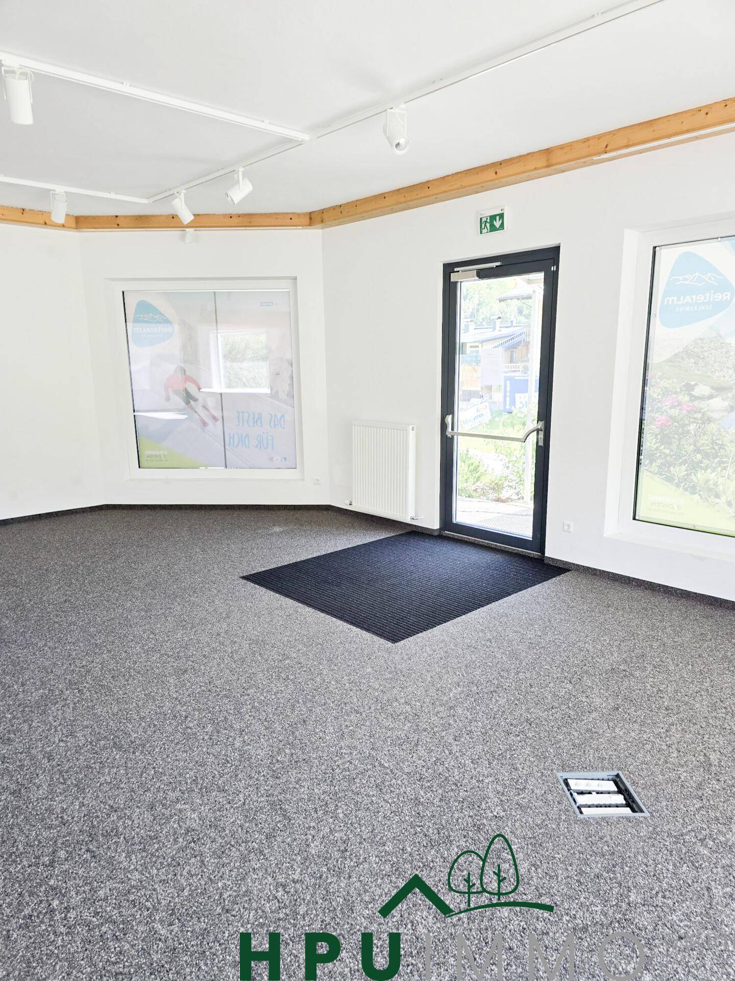 Heller Gewerberaum mit großen Schaufenstern, grauem Teppichboden und moderner Beleuchtung.