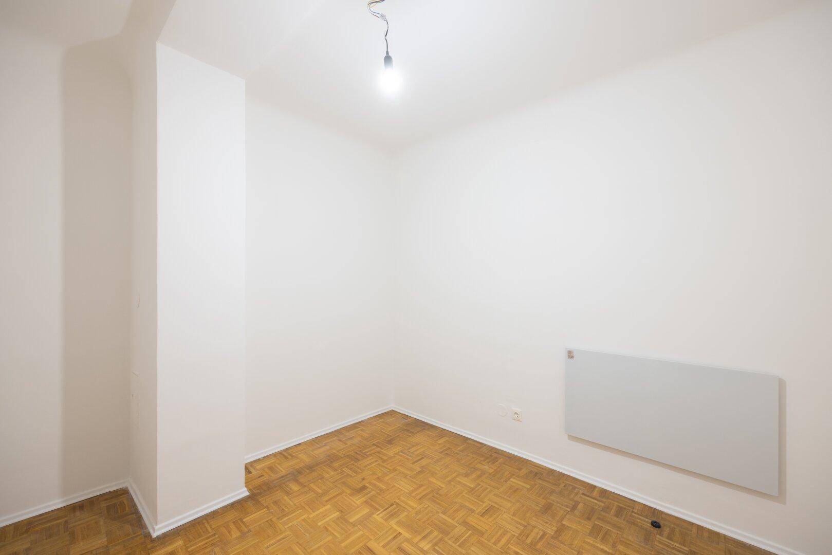 Leeres Zimmer mit Parkettboden, weißen Wänden und einem Heizkörper unter dem Fenster.