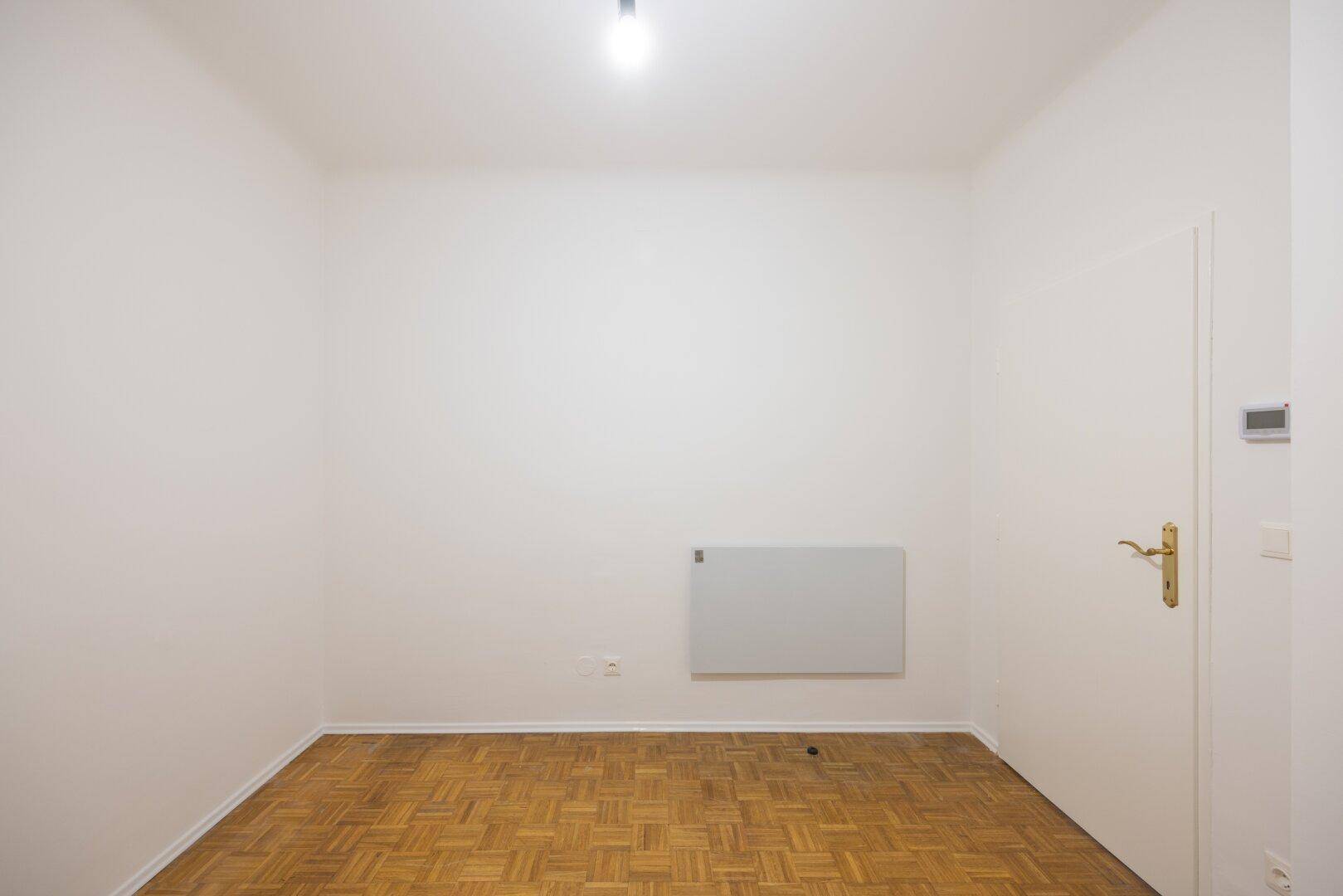 Helles, leeres Zimmer mit Parkettboden, weißen Wänden und einer Tür.