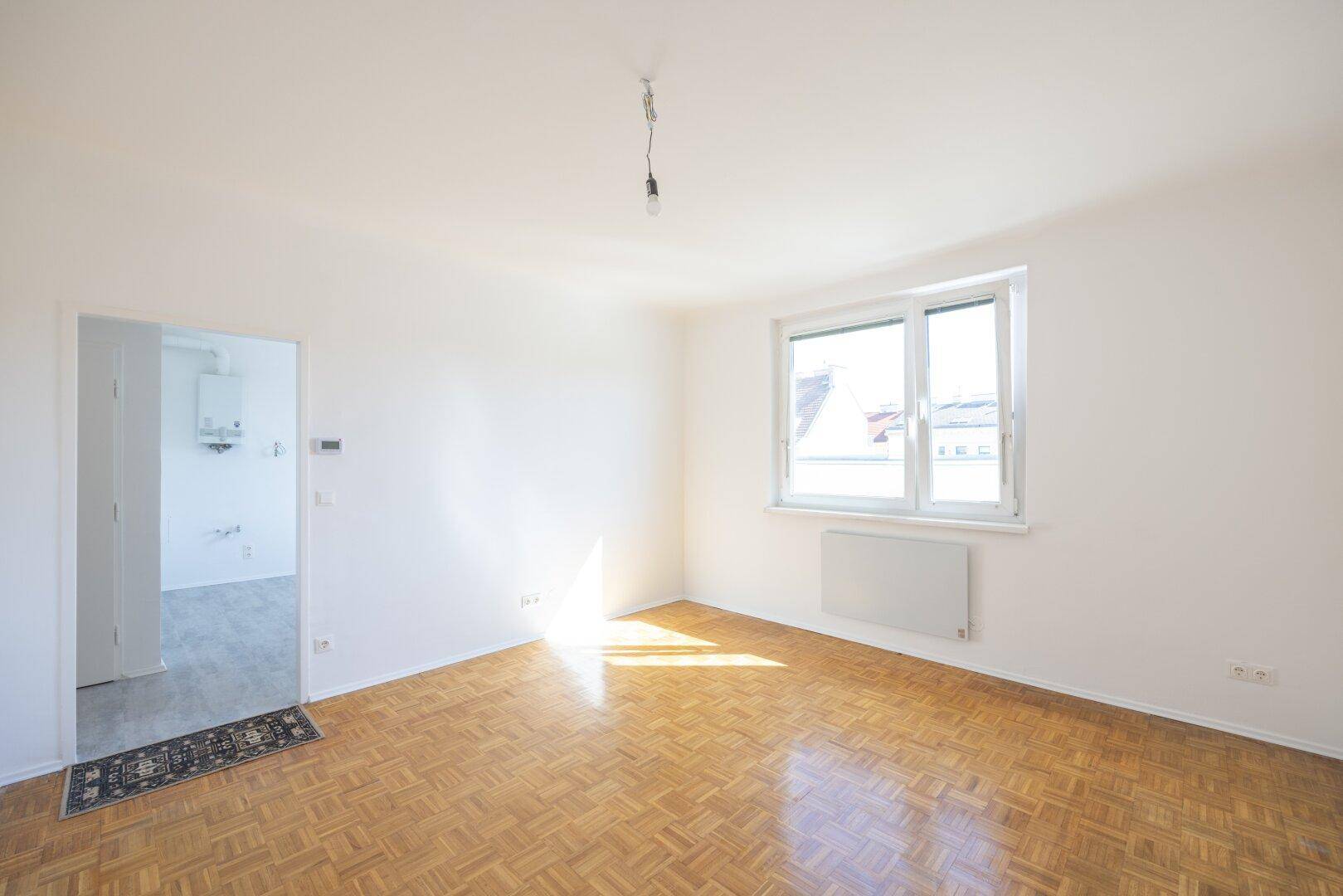 Heller, leerer Wohnraum mit Fischgrätparkettboden und großem Fenster, das viel Sonnenlicht hereinlässt.