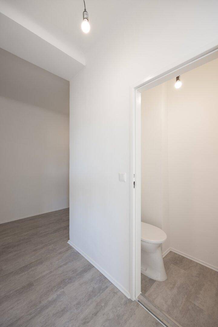 Blick von einem Raum mit grauem Bodenbelag in eine separate Toilette mit WC-Becken.